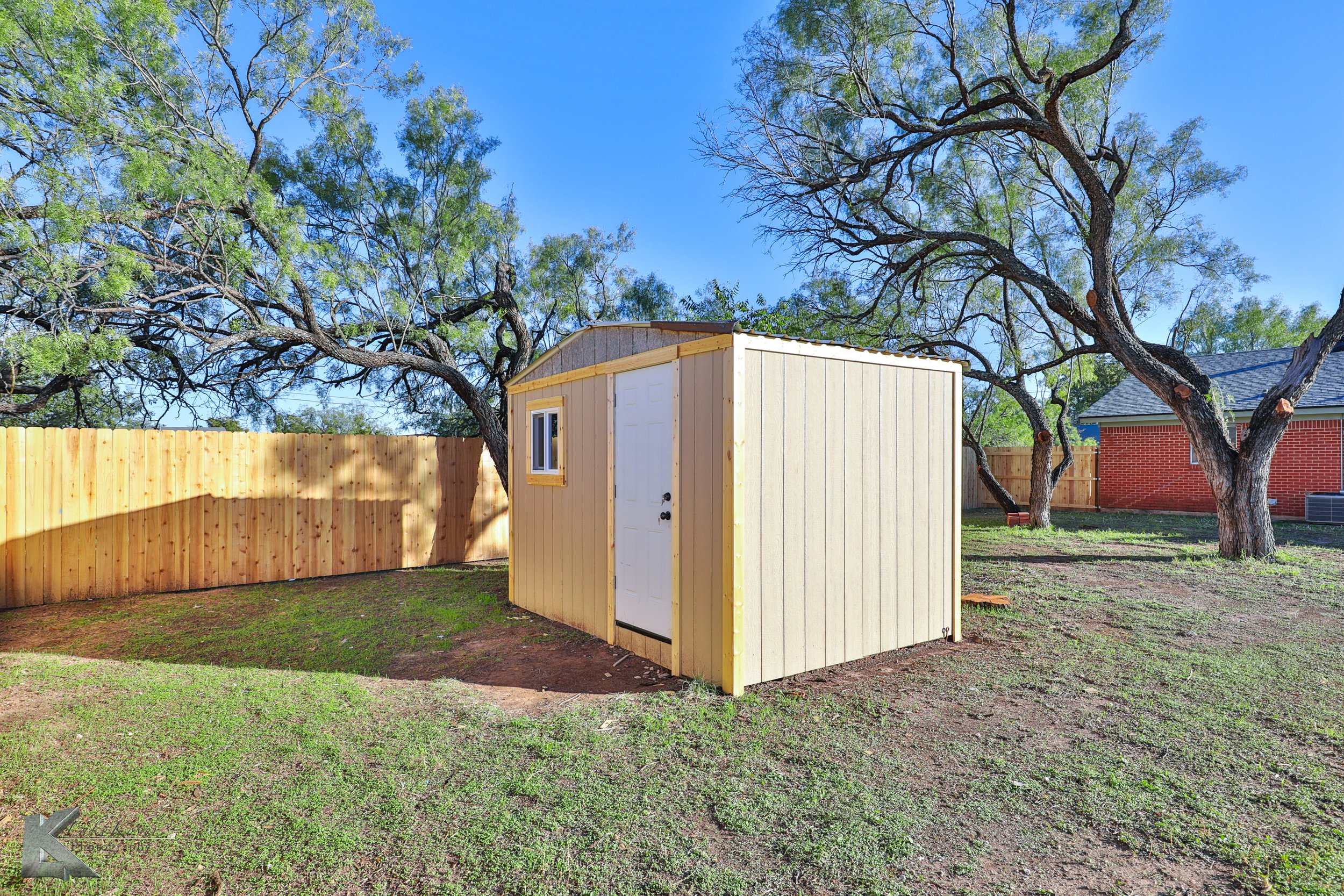 A small yellow shed with a white door and window in a backyard, surrounded by a wooden fence and large trees with green leaves under a clear blue sky.