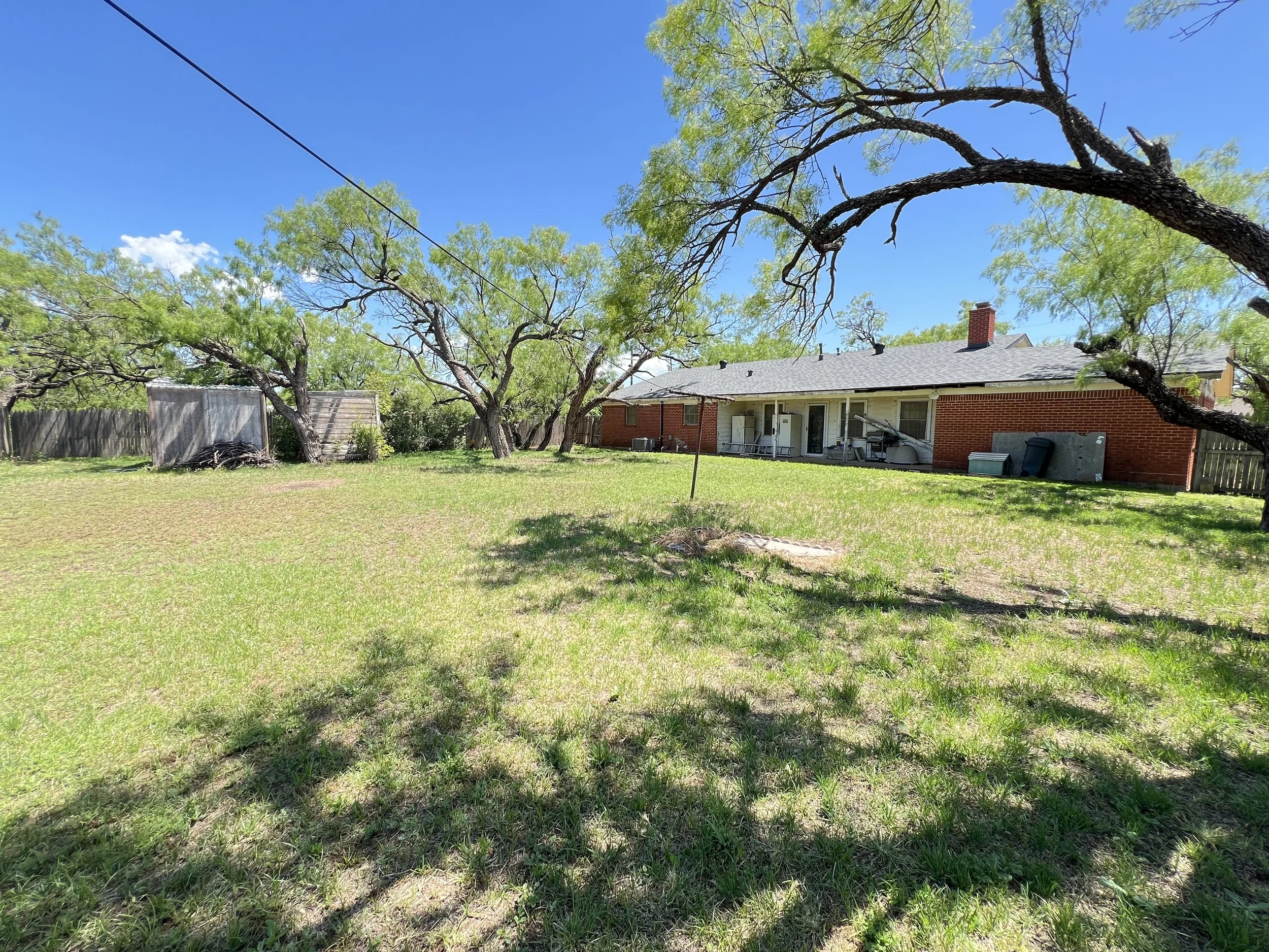 Backyard with green grass, shade trees, and a one-story red brick house with a patio and sliding glass doors.