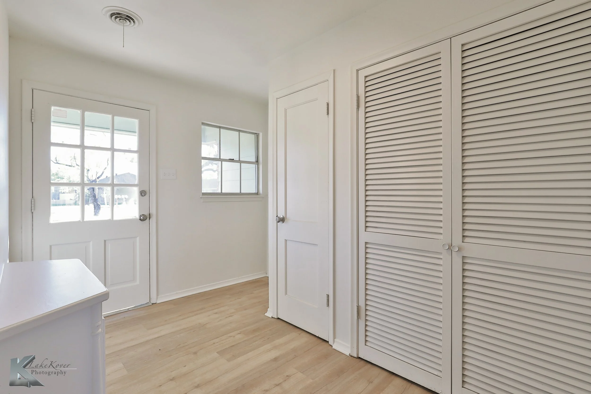 Empty white room with a door, a window, a closet with louvered doors, and light wood flooring.