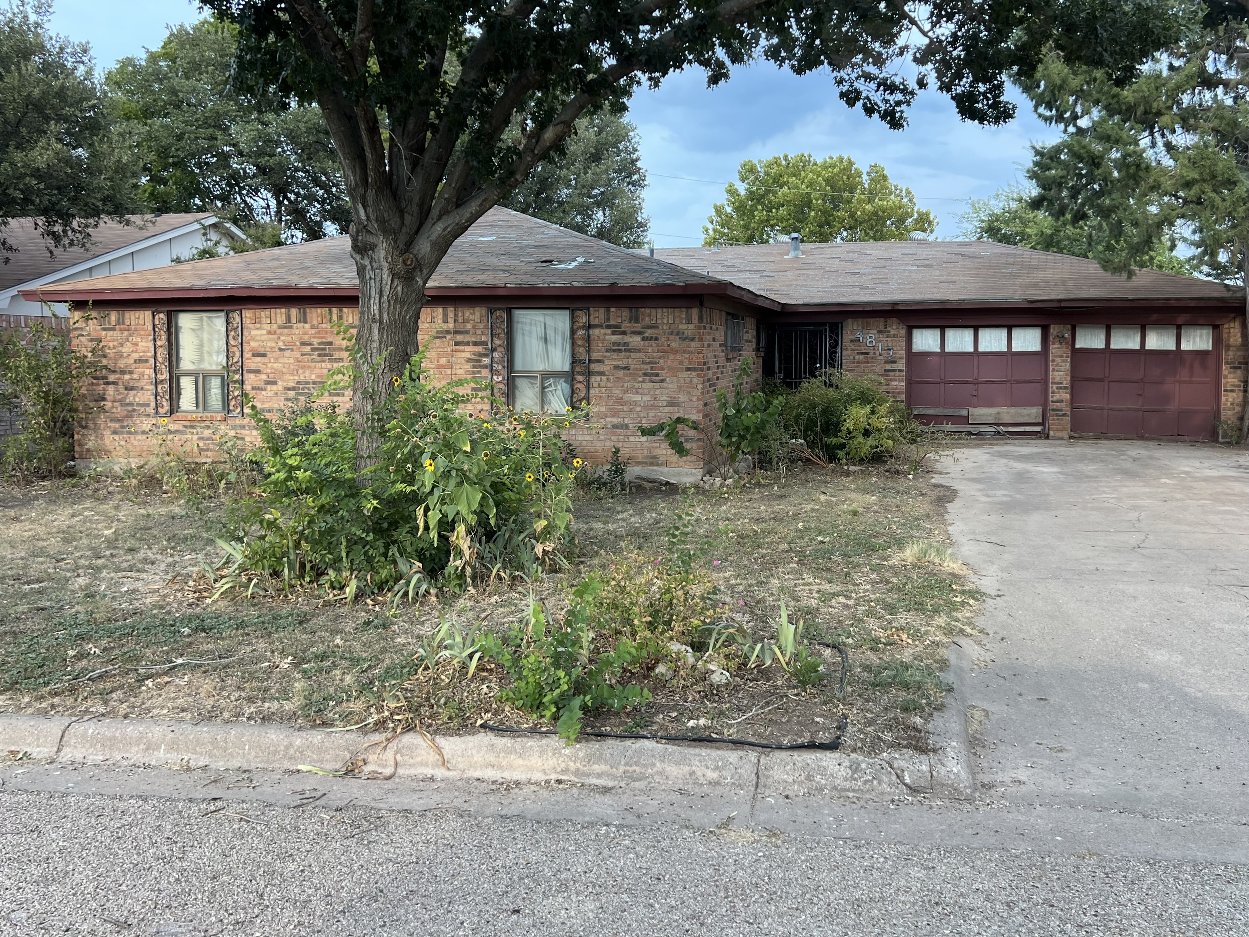 A single-story brick house with a front lawn, large tree, and a driveway leading to a double garage with maroon doors. The house has two front windows with decorative wrought iron bars, and the yard has some plants, bushes, and patchy grass.