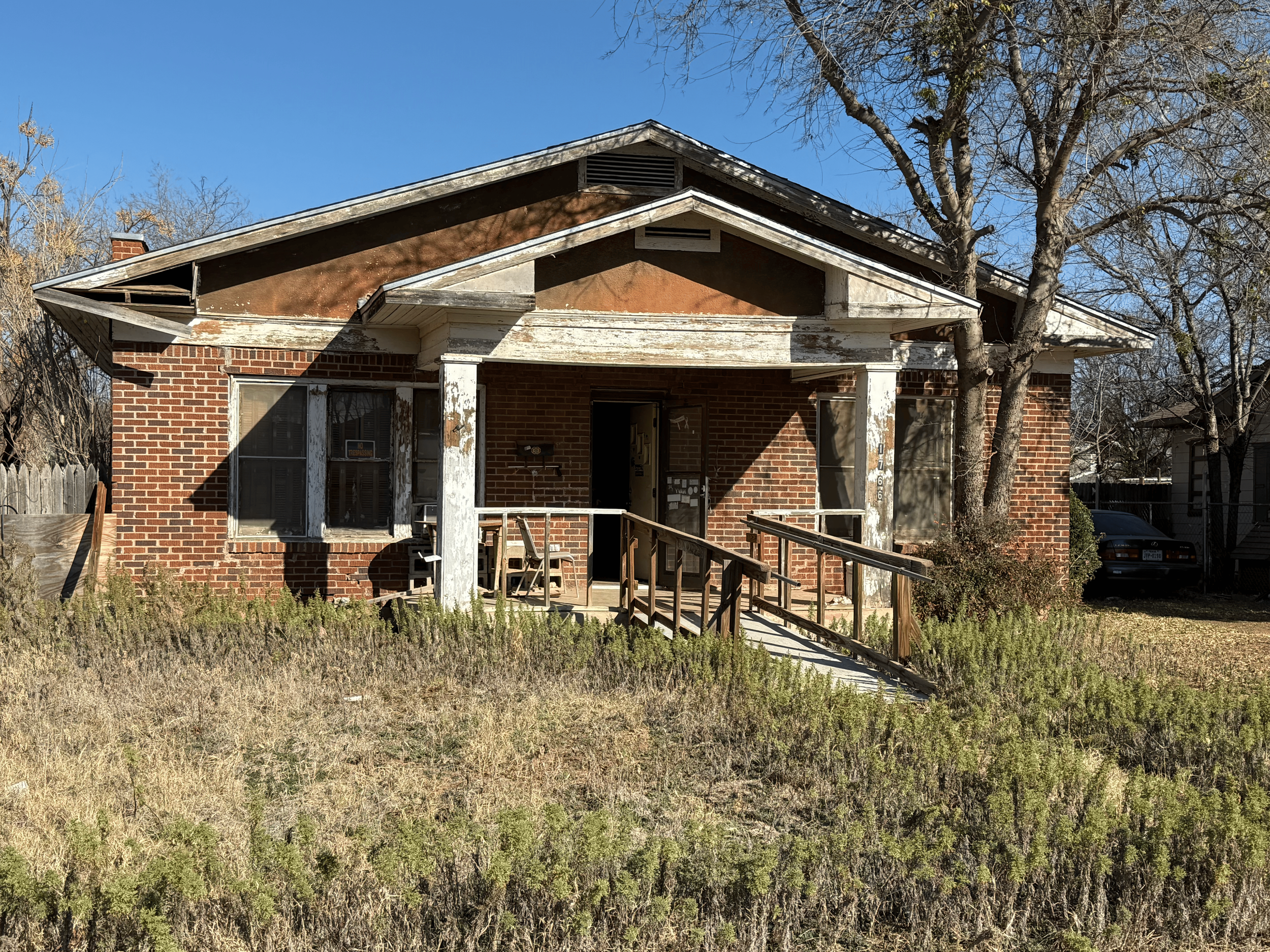 An old, abandoned brick house with peeling white paint on the porch columns, a broken railing, and overgrown grass in the yard. The house has a wide front porch, a ramp leading up to the entrance, and large windows. The roof and porch appear weathered.