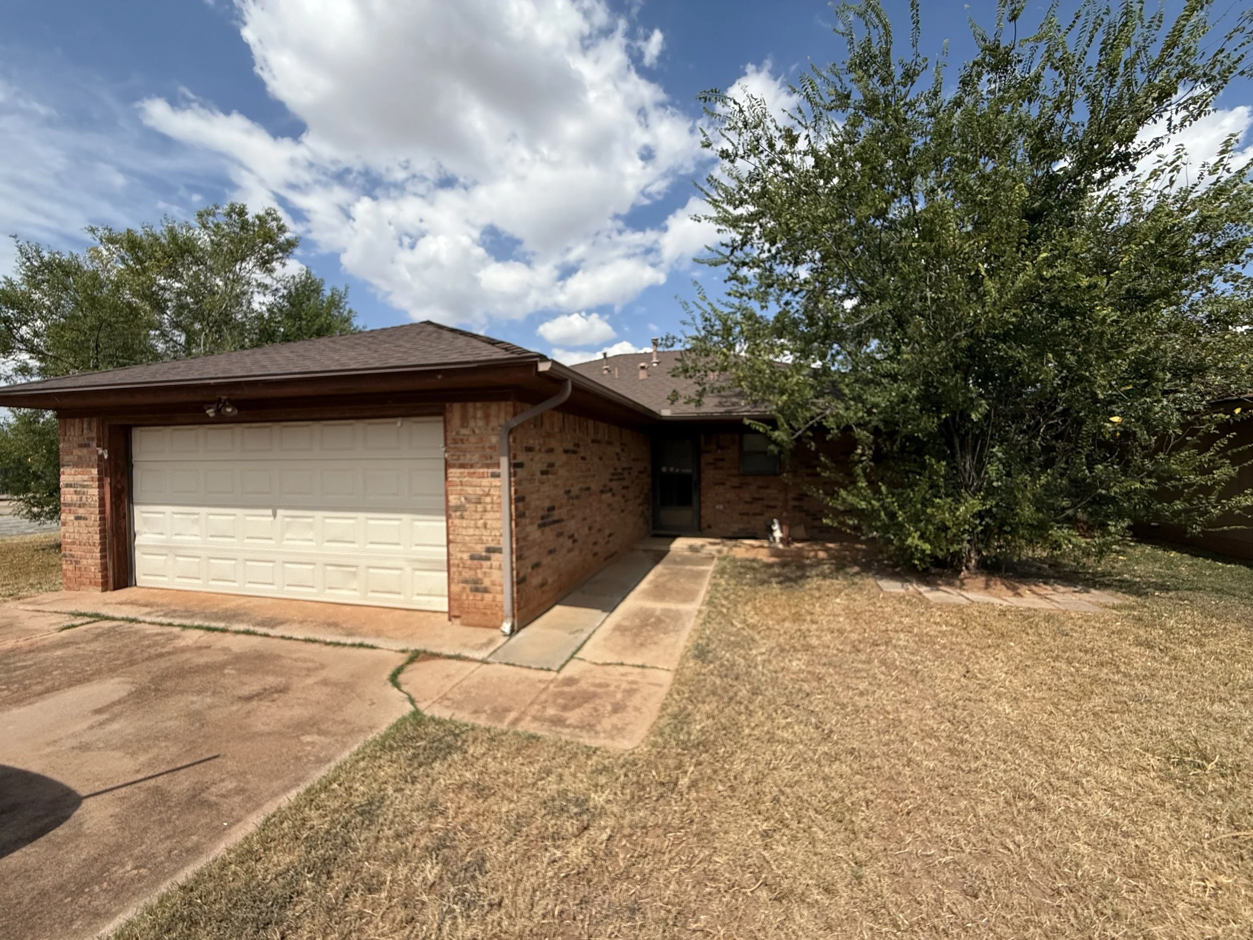 A single-story brick house with a garage door, concrete driveway, front porch, and large tree next to the house, under a partly cloudy sky.