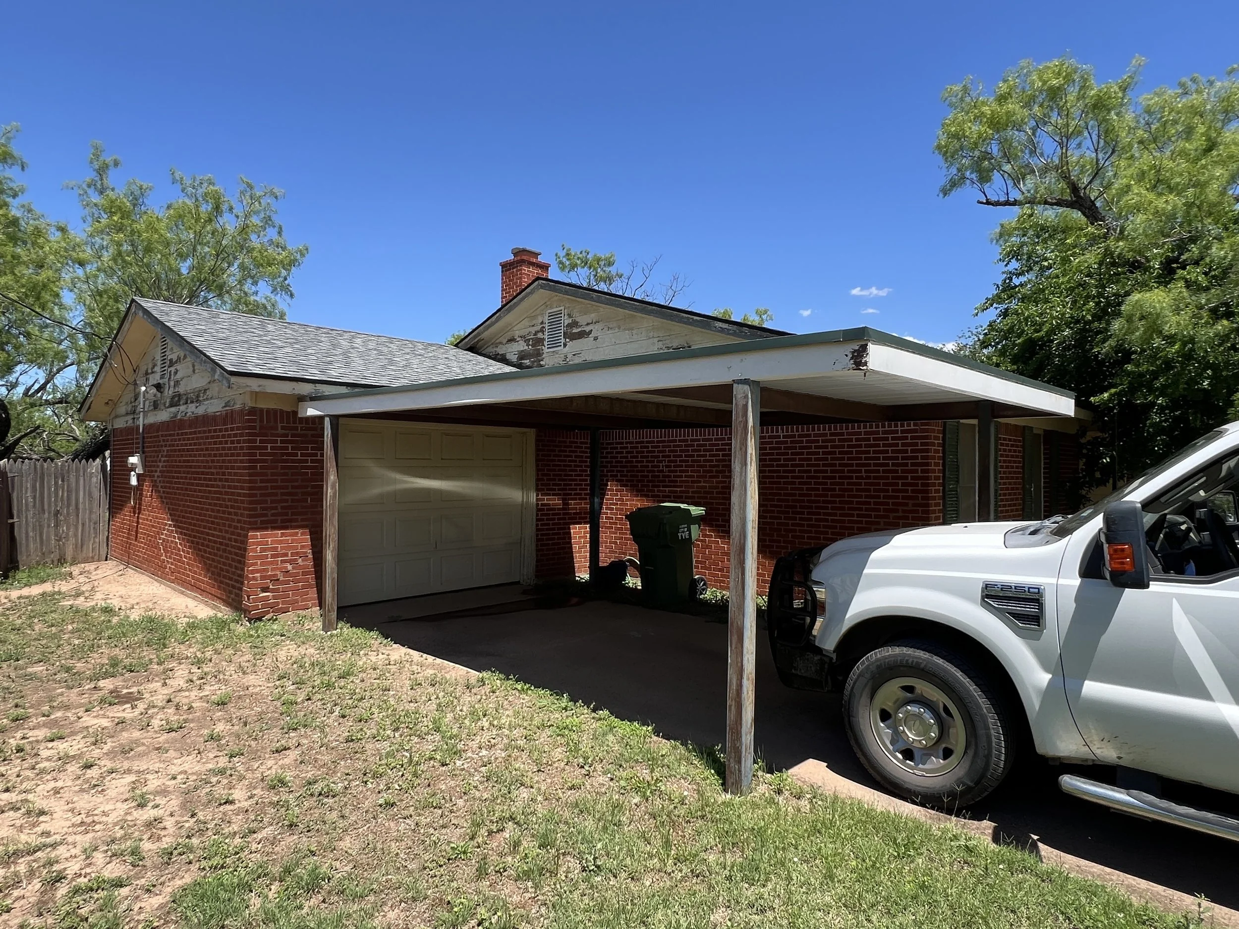 A house with a brick exterior, an attached carport, a white Ford F-250 truck parked underneath, a green trash bin, and green trees in the background on a sunny day.