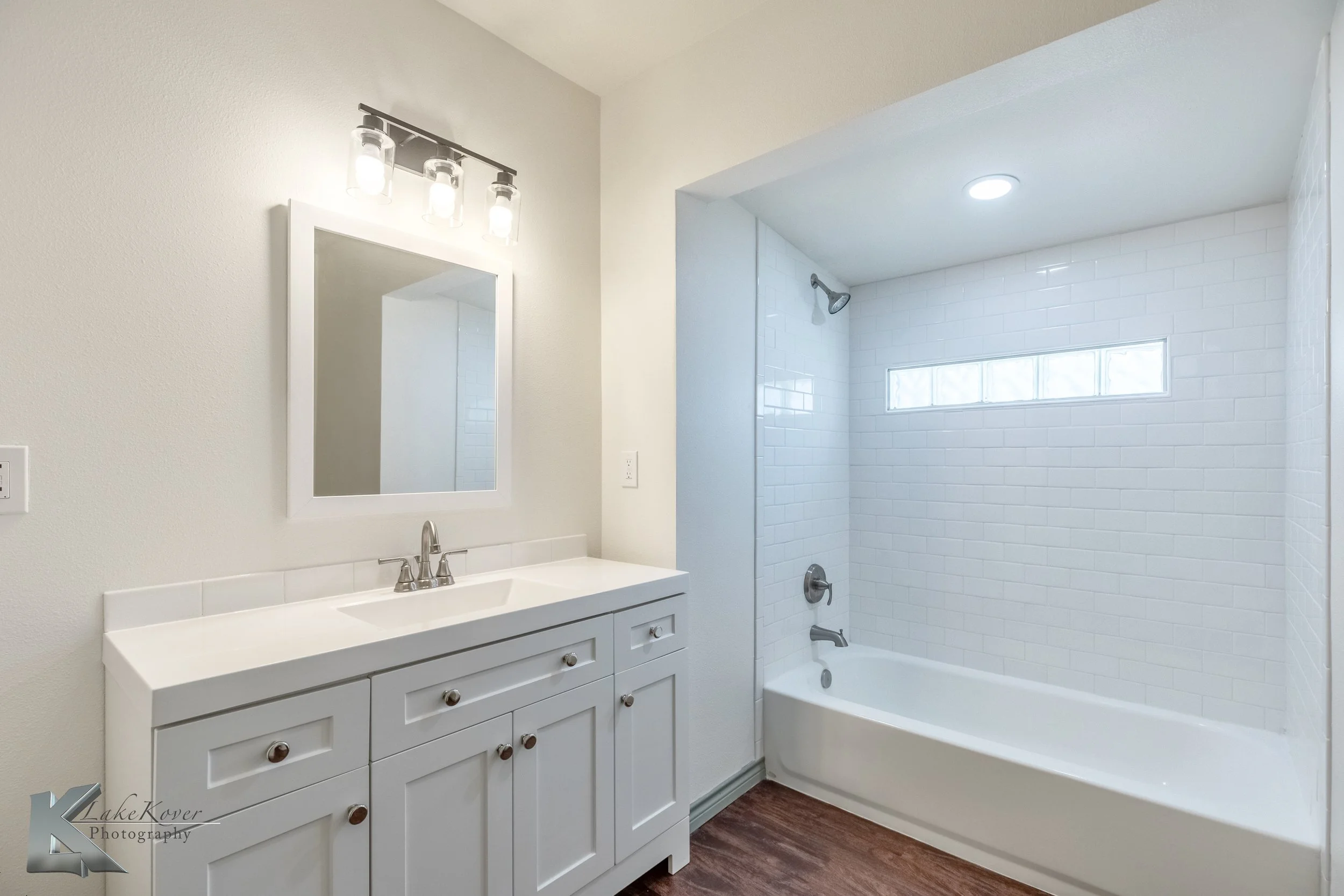 Modern bathroom with white vanity, mirror, and a bathtub with white subway tile surround and a horizontal glass block window.