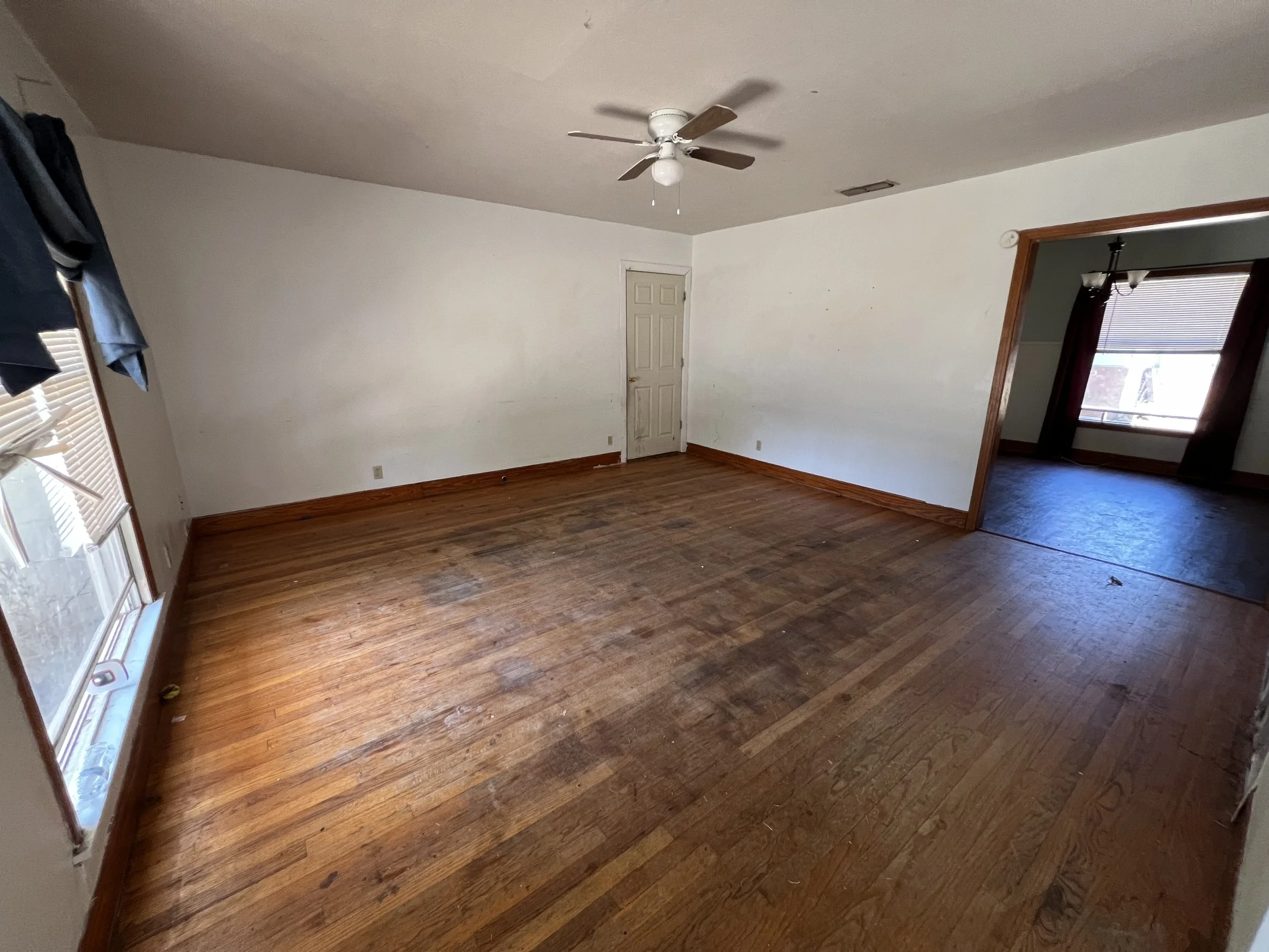 Empty room with hardwood floors, white walls, a ceiling fan, and a window with blinds and curtains.