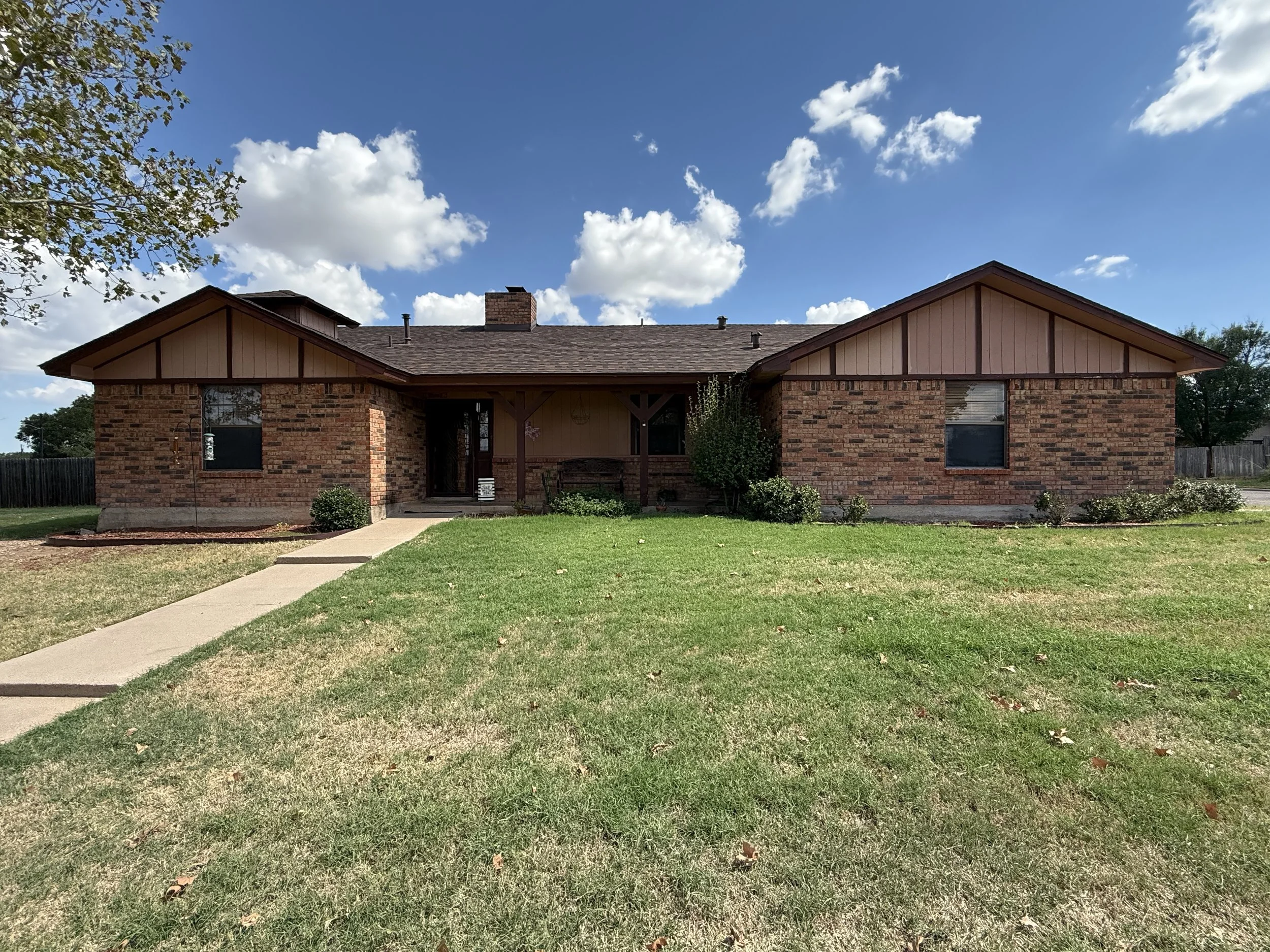 A single-story brick house with a front porch, surrounded by a grassy lawn and some bushes, under a partly cloudy blue sky.