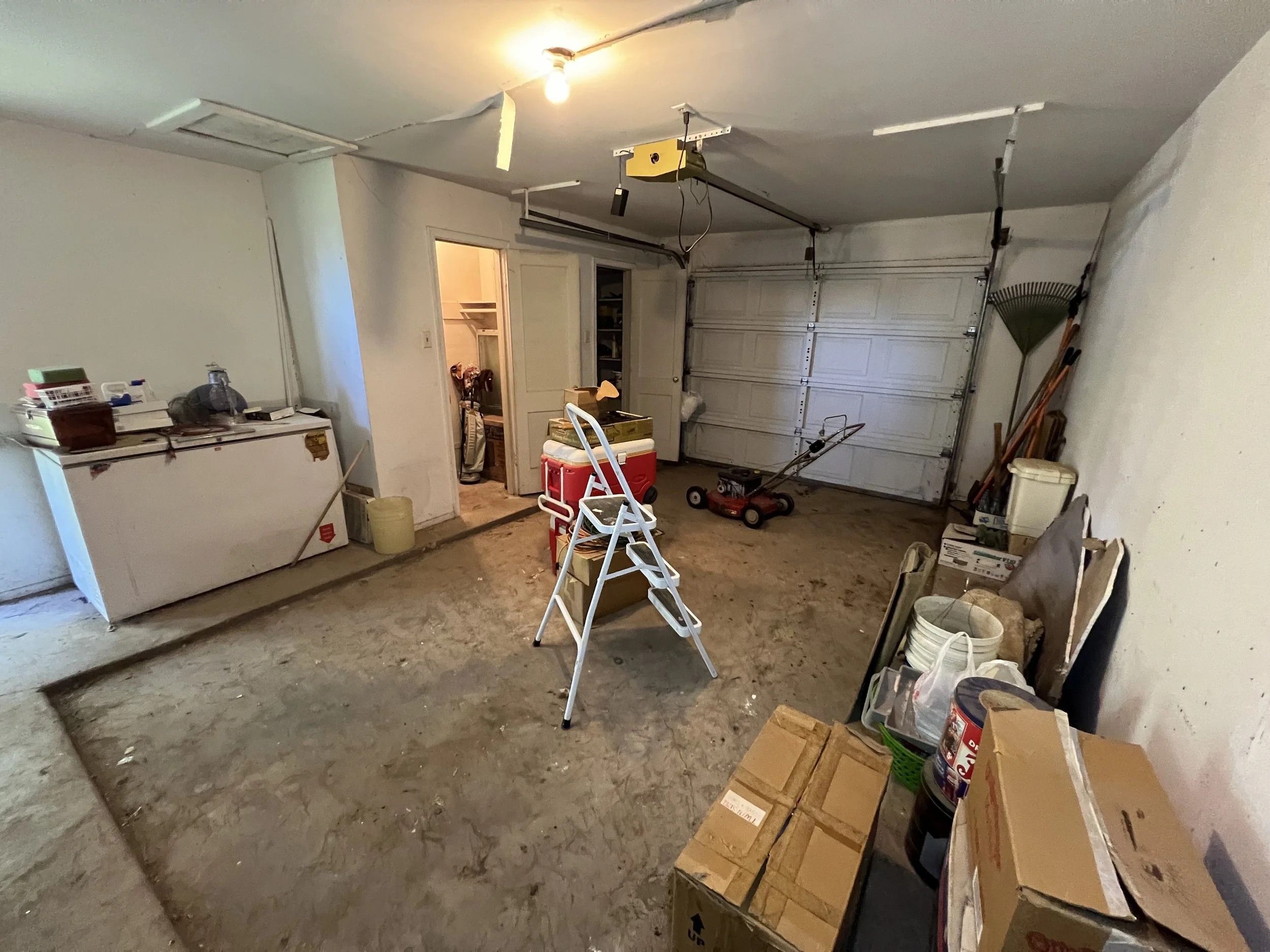 A cluttered garage with a partially finished concrete floor, a closed white garage door, and various tools, boxes, and storage items scattered throughout. There is an open doorway leading to a storage room or closet, with a ladder and a cooler in fro