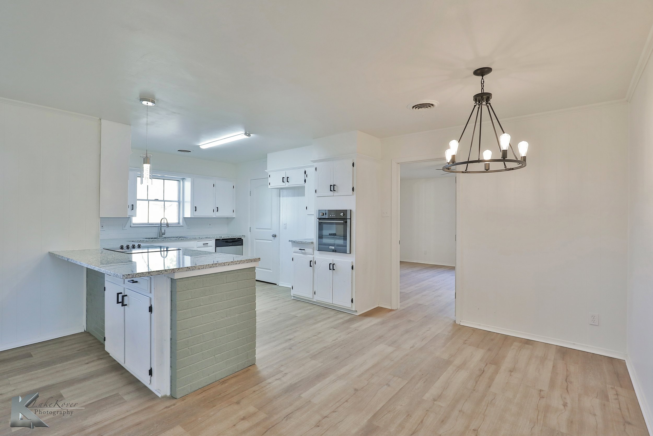 Empty kitchen and dining area with white cabinets, granite countertops, light wooden flooring, a black chandelier, and a window in the kitchen.