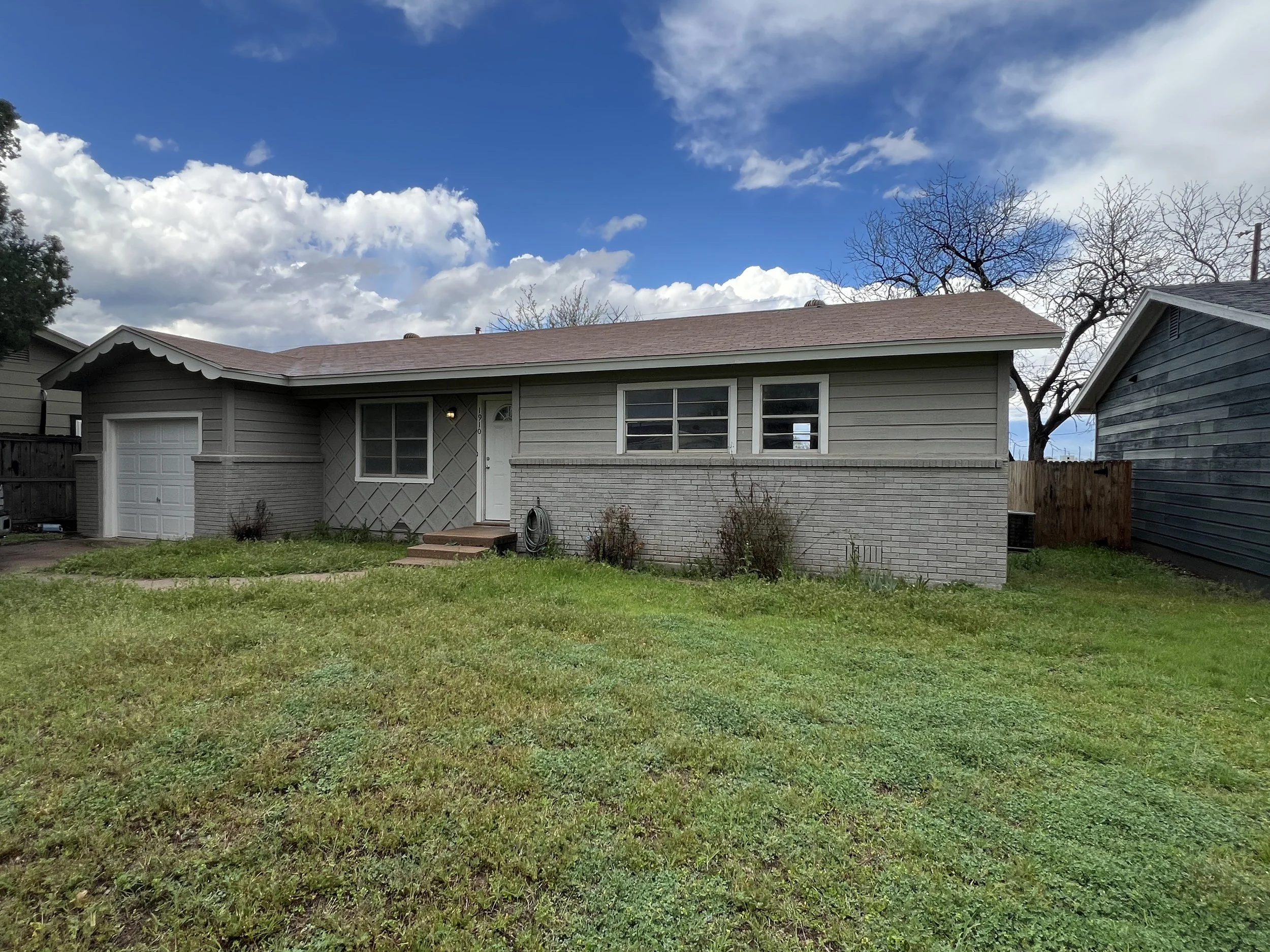 A single-story house with gray siding and a brick lower section, a one-car garage, a small front porch with steps, a grassy yard, and a blue sky with clouds.
