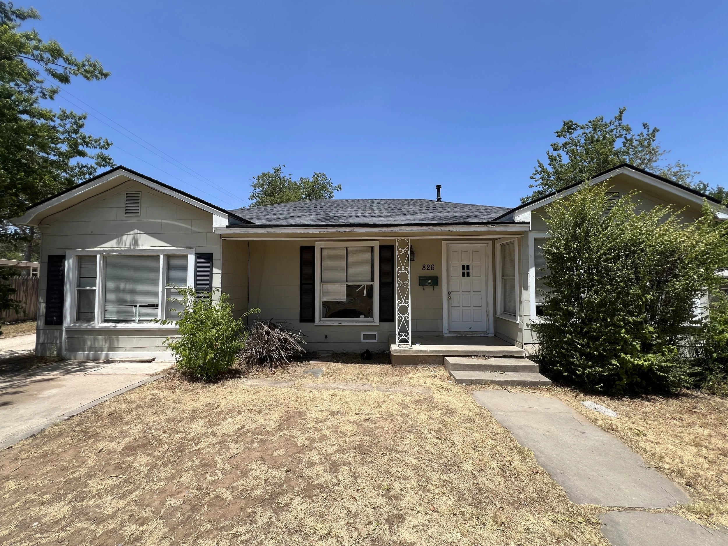 Front view of a single-story house with a small front porch, steps, and sidewalk. The house has a light exterior, black shutters, and some bushes and trees in the yard under a clear blue sky.