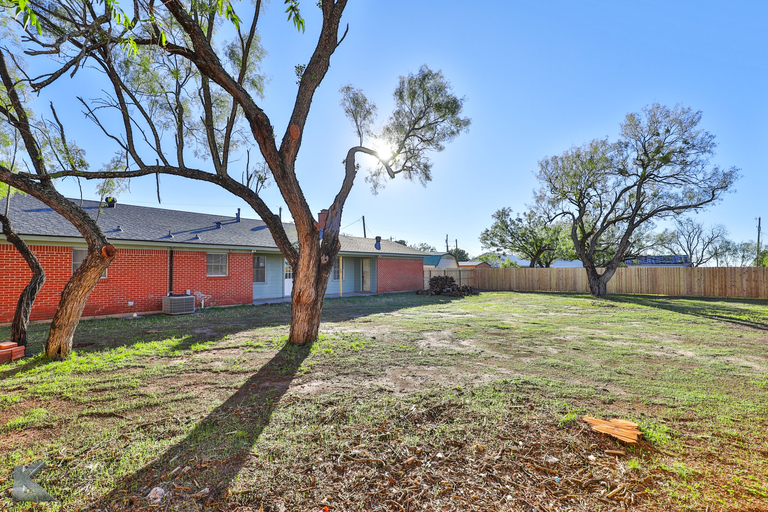 Backyard with bare grass, a few trees, and a brick house with a sloped roof under a clear blue sky and sunlight.