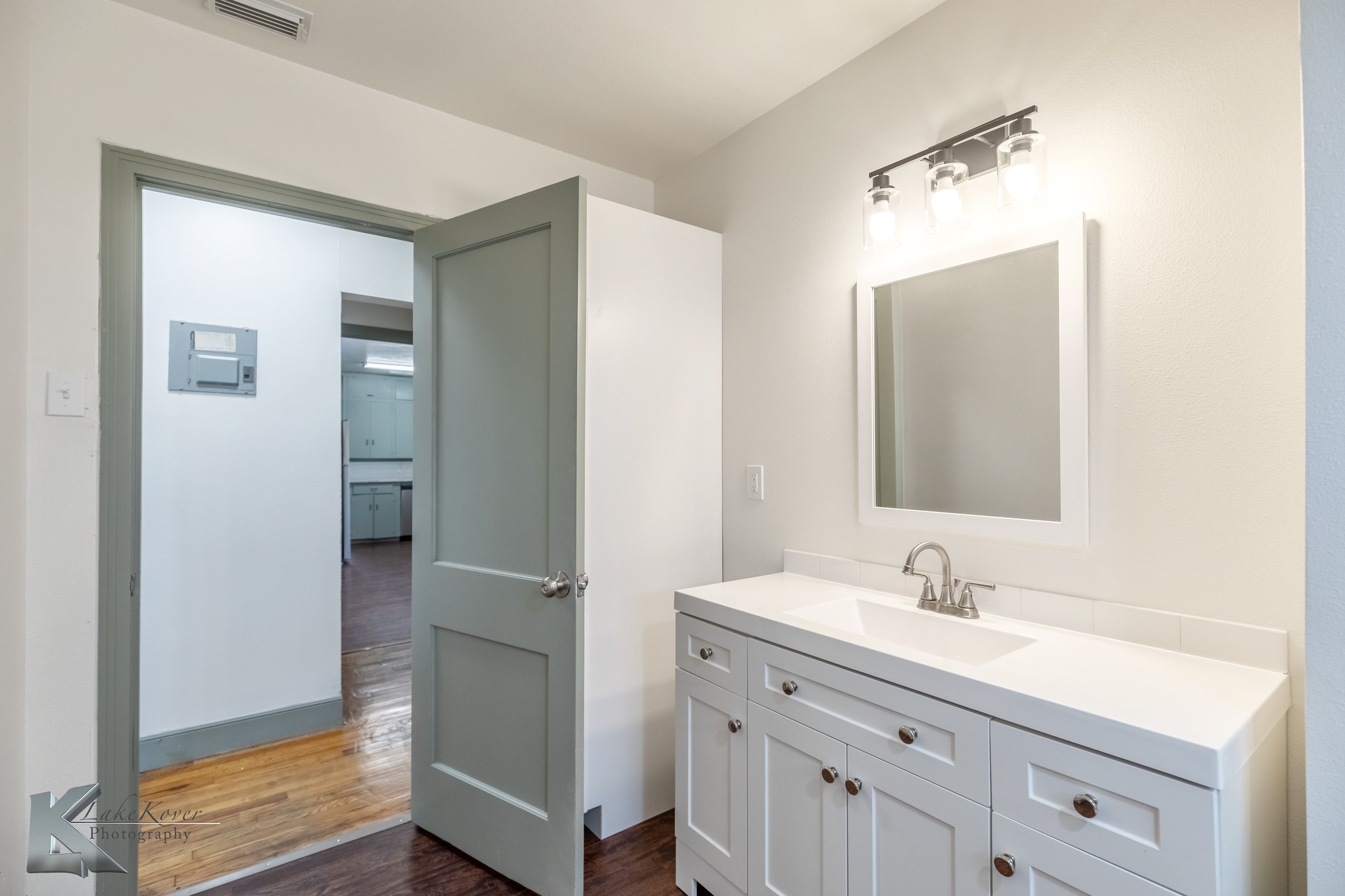 Bathroom with a white vanity, mirror, and light fixture, doorway leading to another room, hardwood floor.