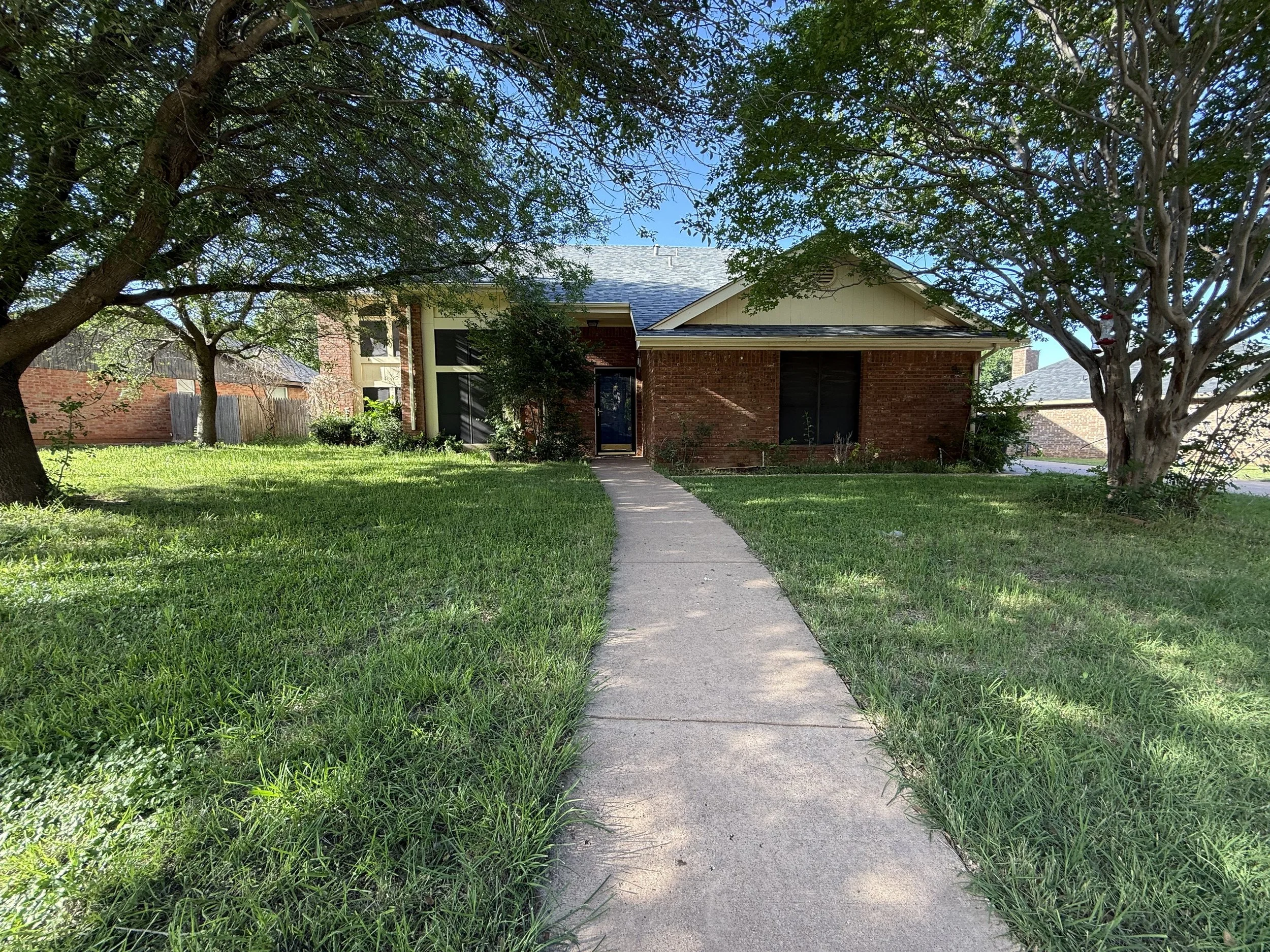 Front view of a brick house with a sidewalk leading to the entrance, surrounded by green grass and large trees.