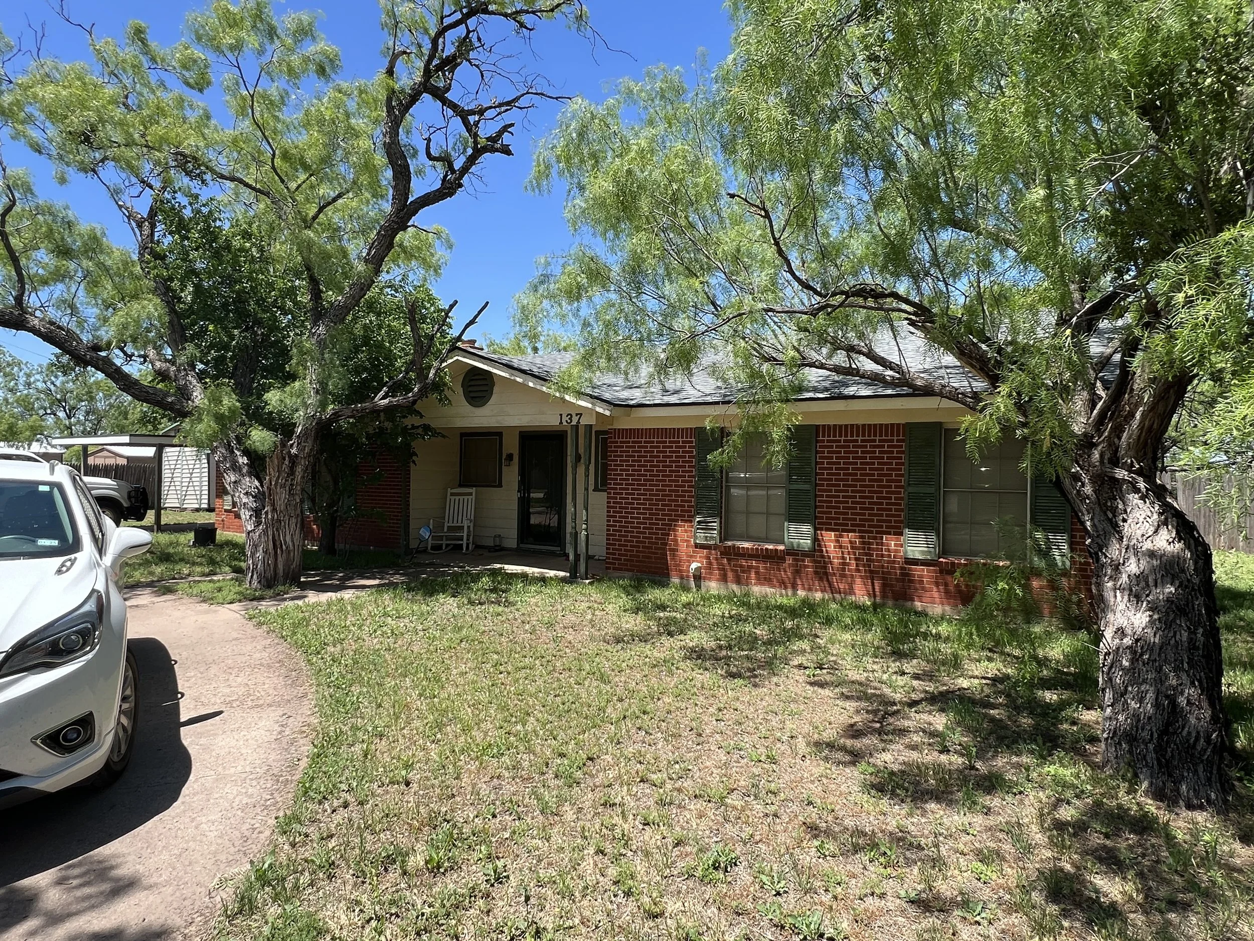 Front yard of a single-story brick and siding house with green shutters, two large trees and a driveway with a white car parked on it, under a clear blue sky.