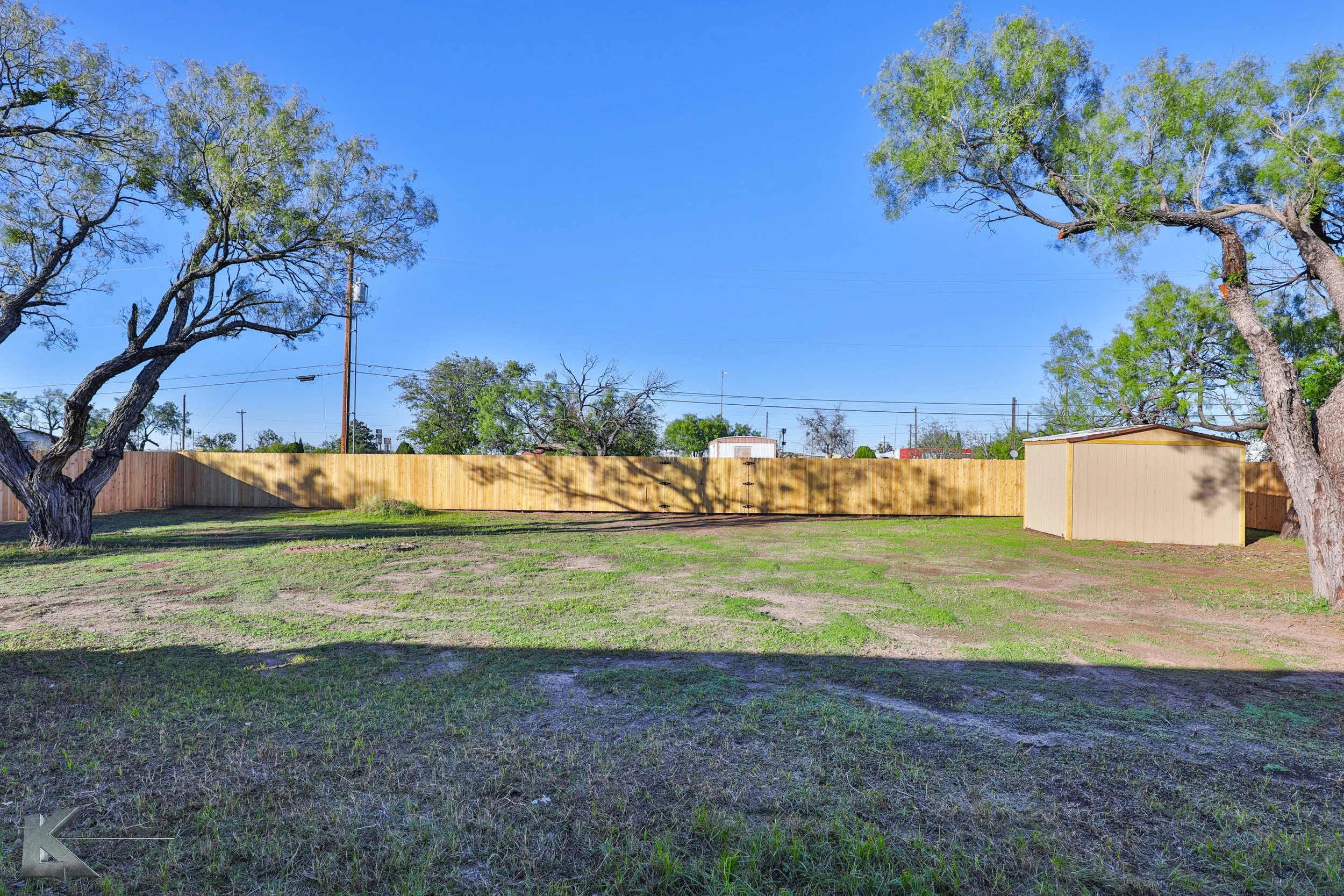 A large backyard with a wooden fence, two large trees, and a small beige shed under a clear blue sky.