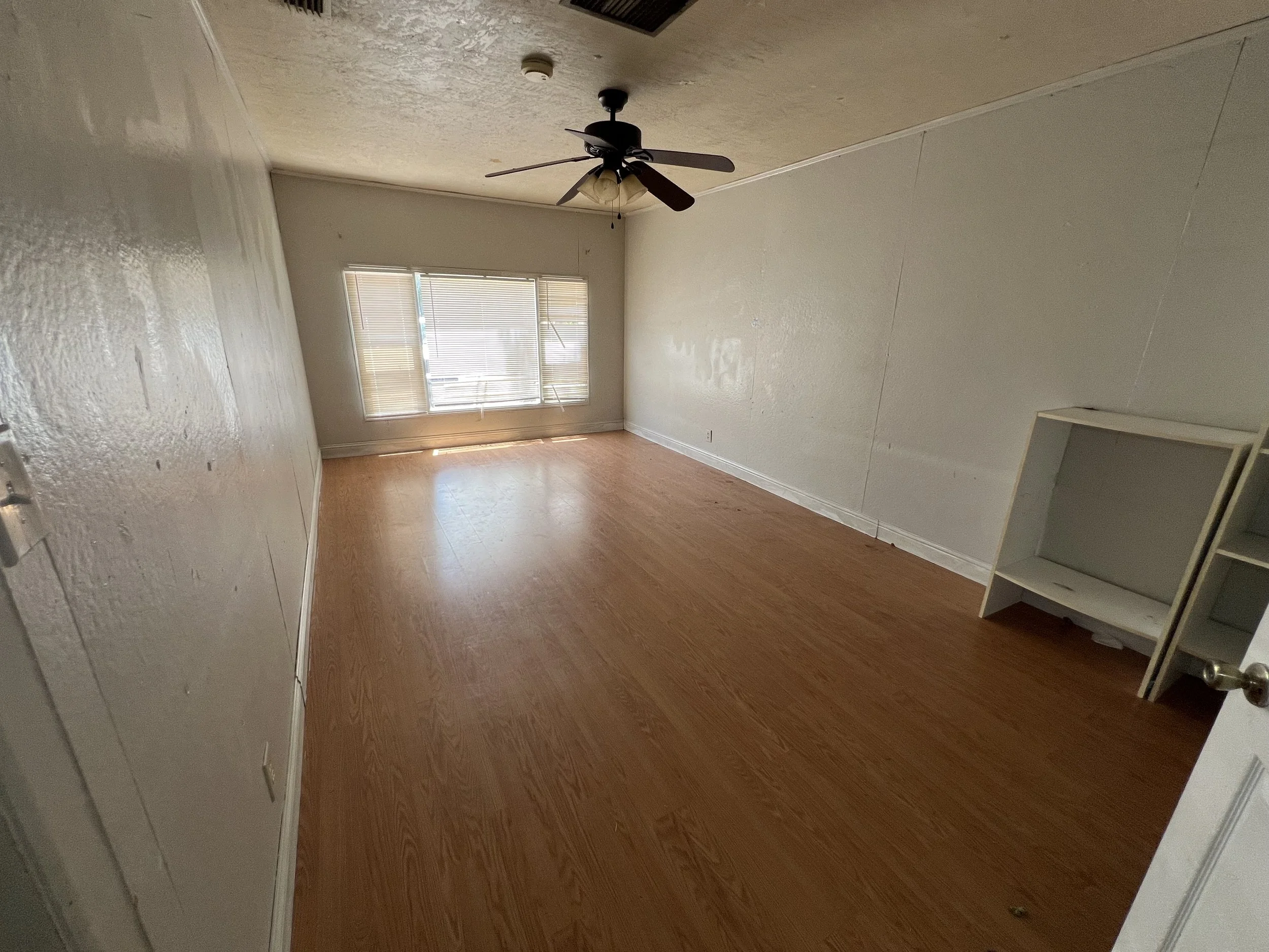 An empty rectangular room with hardwood floors, white walls, a ceiling fan, and a window with blinds on the far wall.