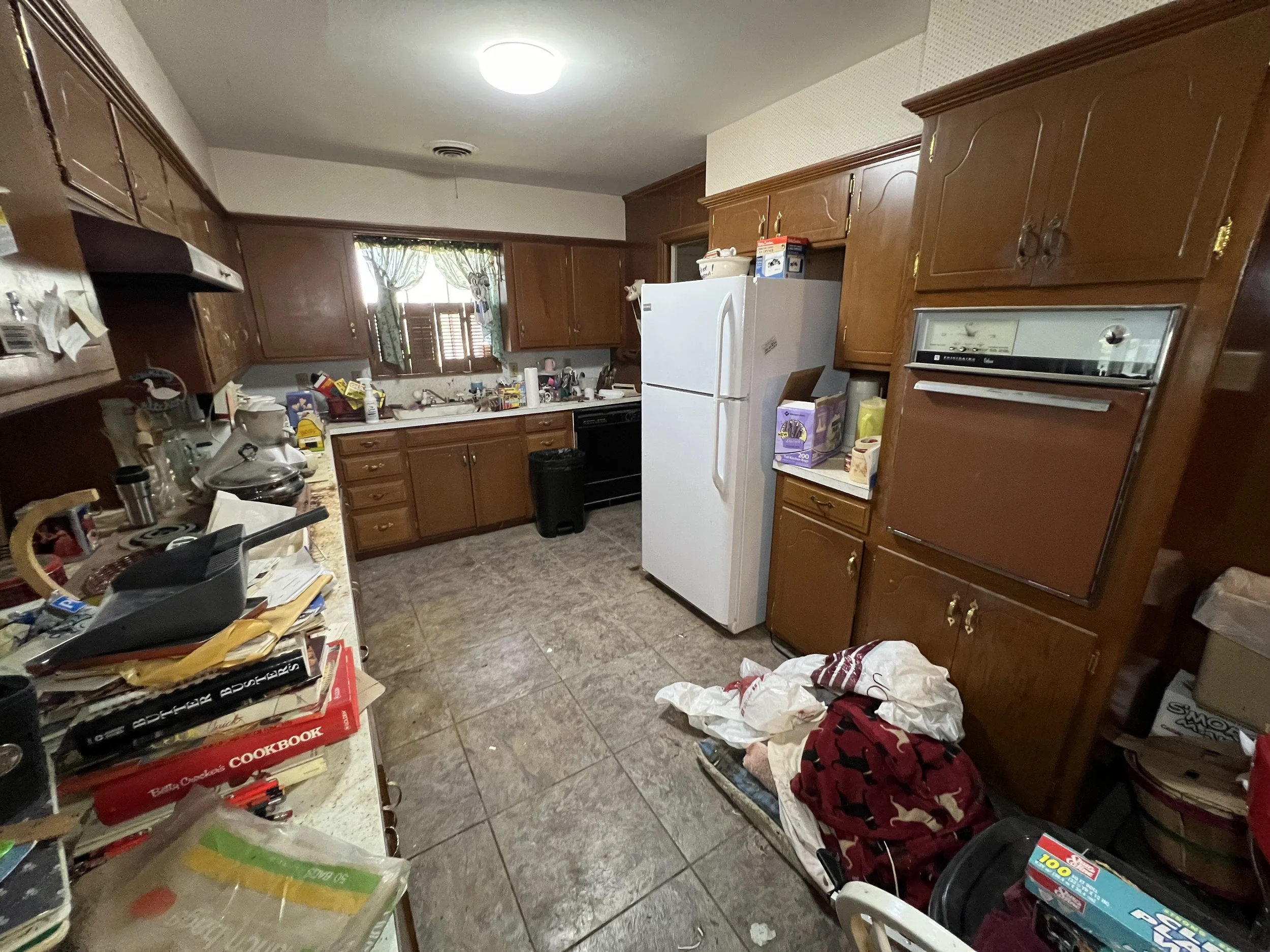 A cluttered kitchen with wooden cabinets, a white refrigerator, a black oven, a window with green curtains, and a countertop with various kitchen items. The floor is tiled and there are miscellaneous items and paper on the countertop and floor.