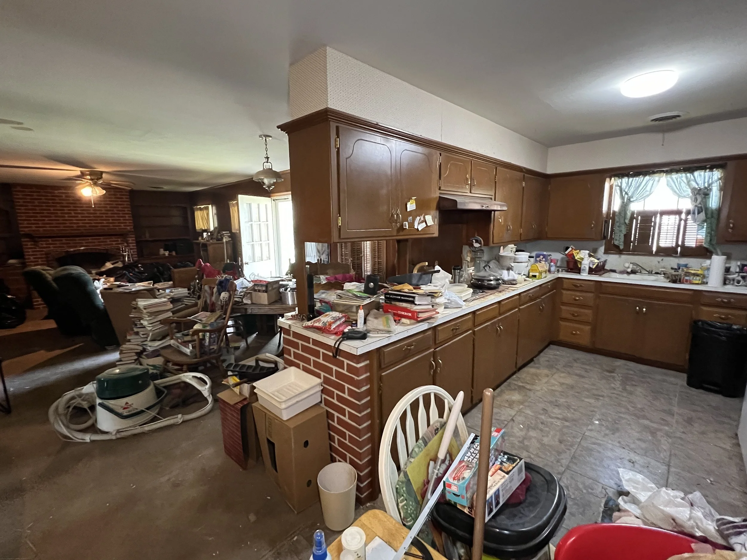 Messy kitchen with cluttered countertops, disorganized dining area with books and papers, and living room with recliner chairs and fireplace in the background.