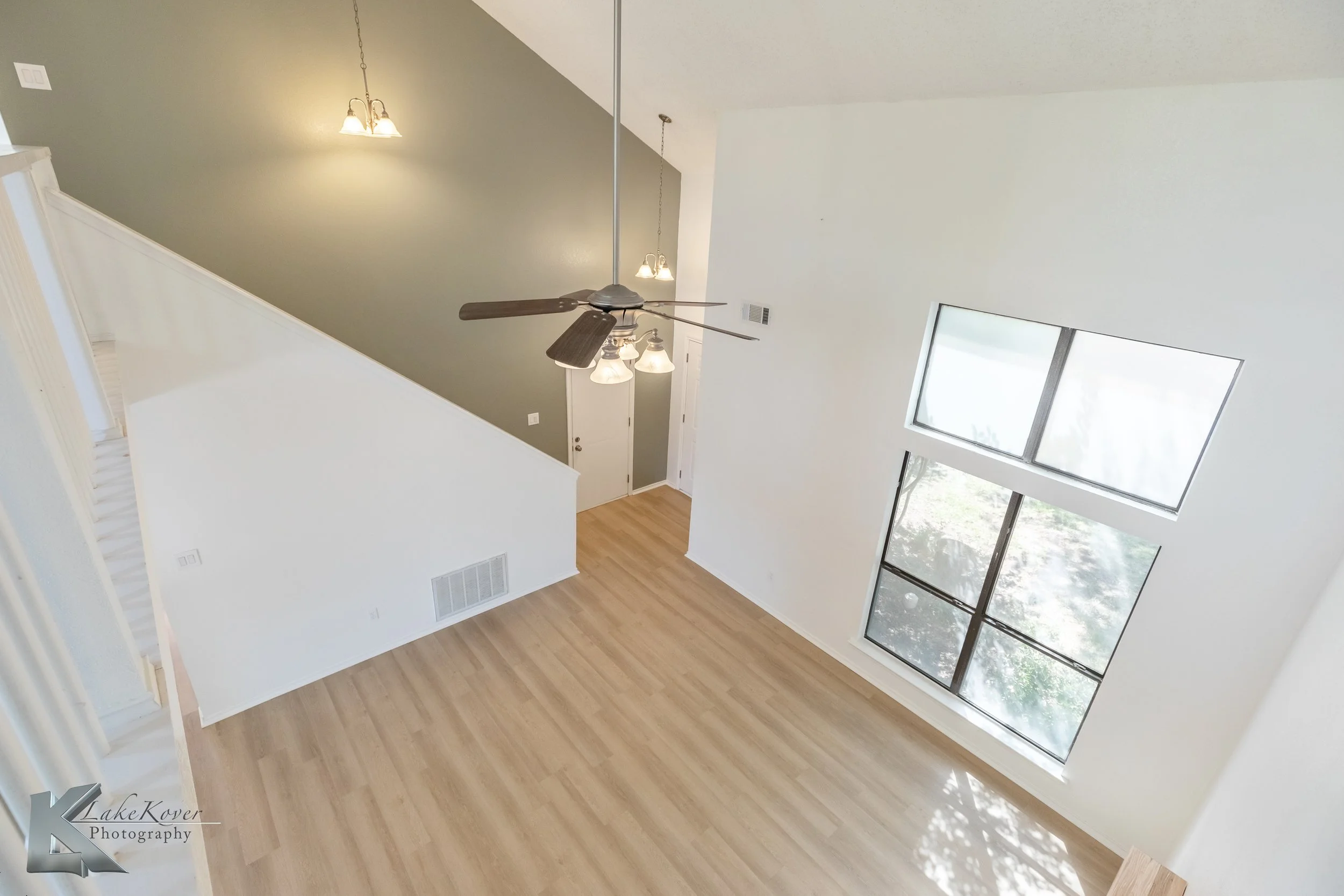 An empty living room with light wood flooring, large windows, a high ceiling, a ceiling fan with lights, and a staircase leading to the upper level.
