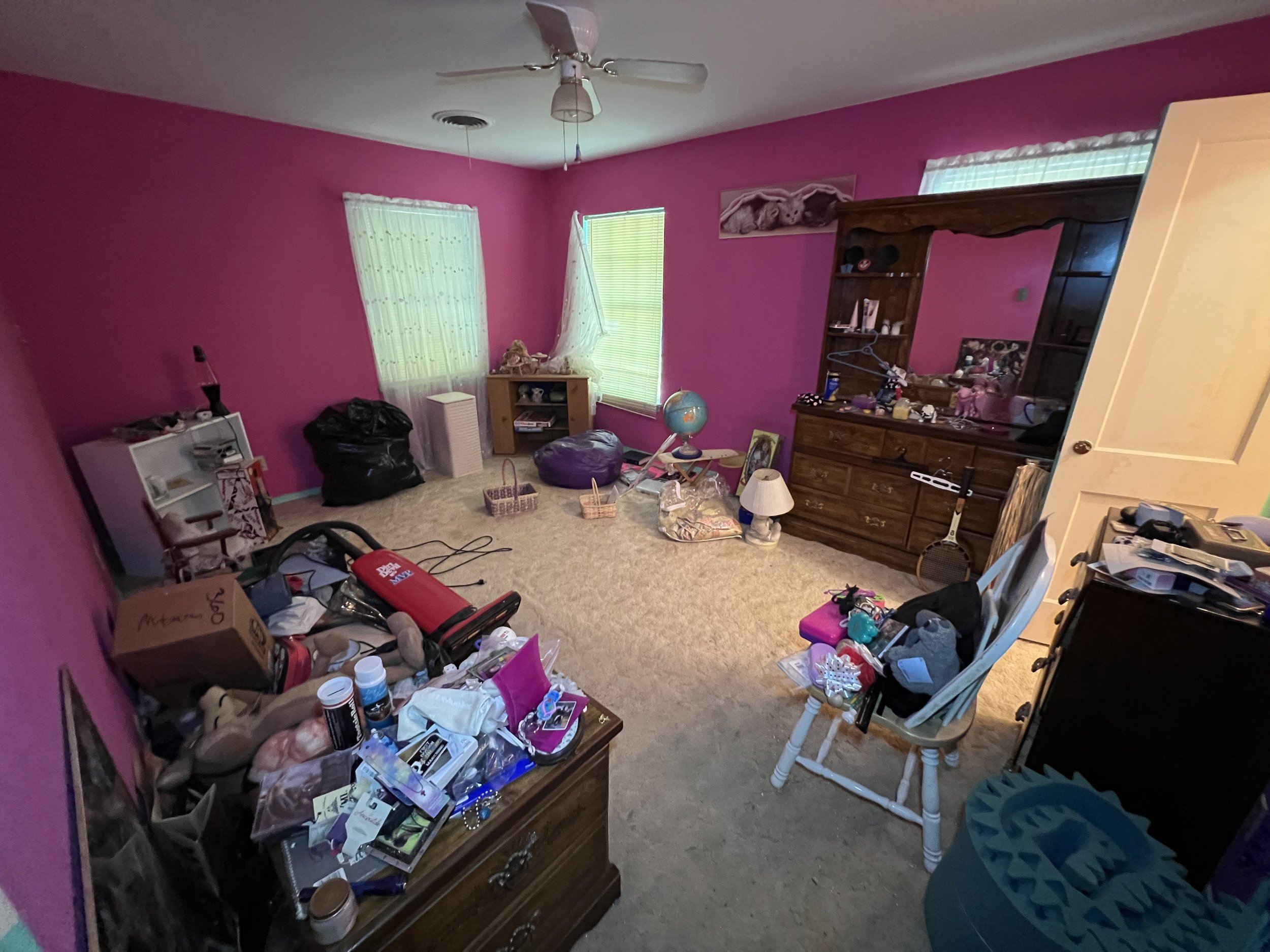 A cluttered bedroom with pink walls, beige carpet, and various furniture and items scattered around, including a dresser with mirror, a small table with a globe, and a white chair with items on it.