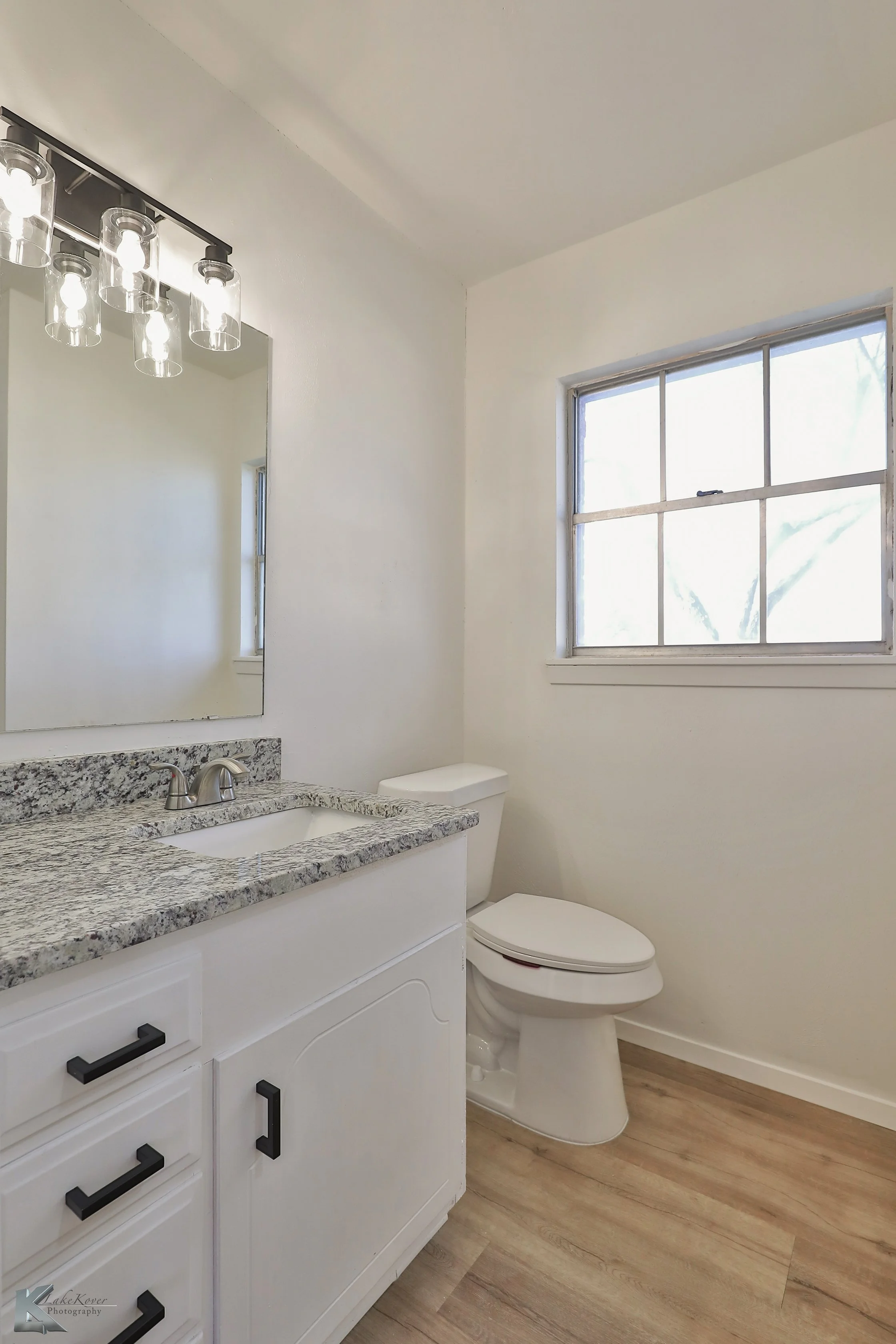 A small bathroom with a white vanity, granite countertop, and black handles, a mirror above, a white toilet, a window with a view of greenery outside, and wood-patterned flooring.