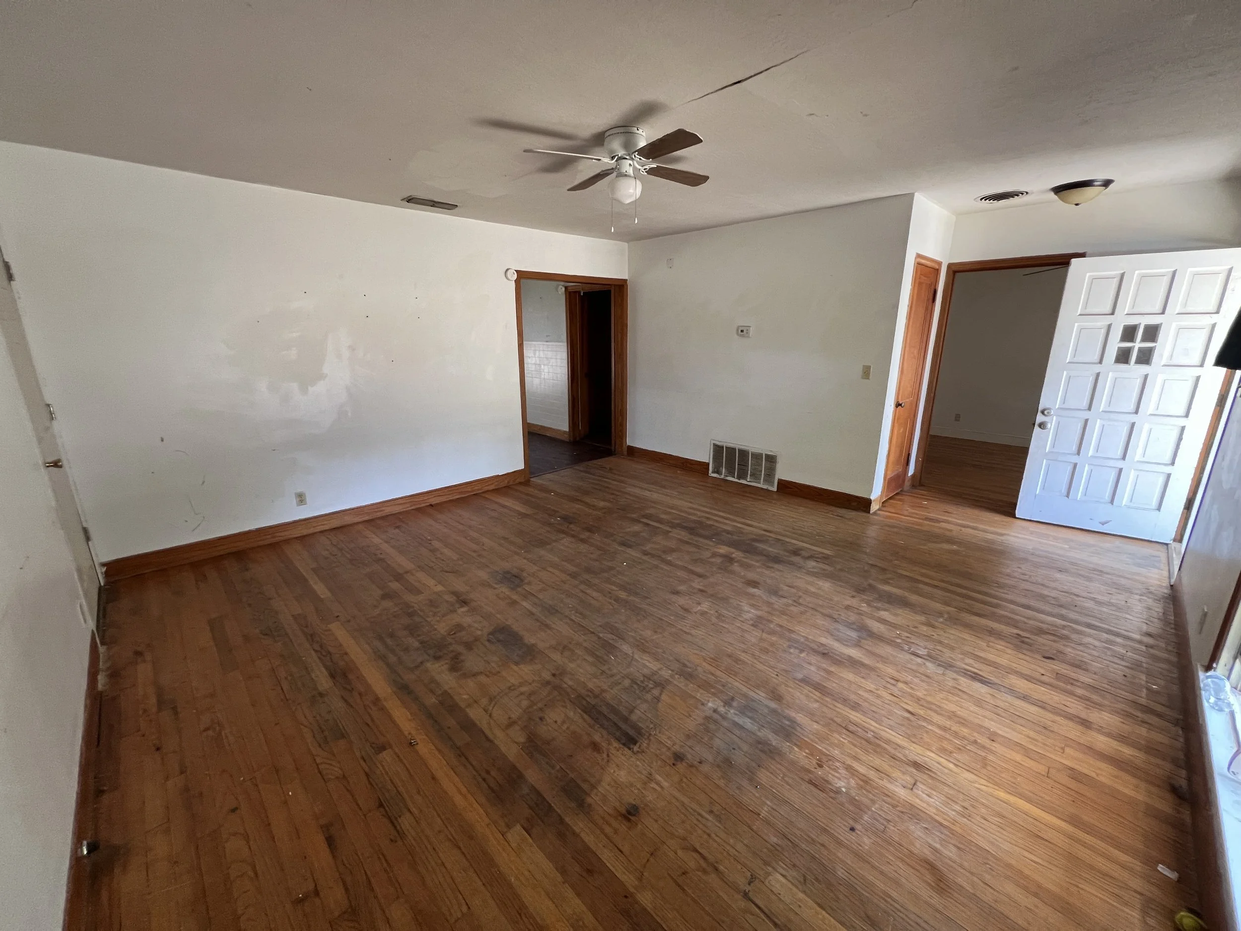 An empty living room with wooden floors, white walls, a ceiling fan, and an open door leading to another room.