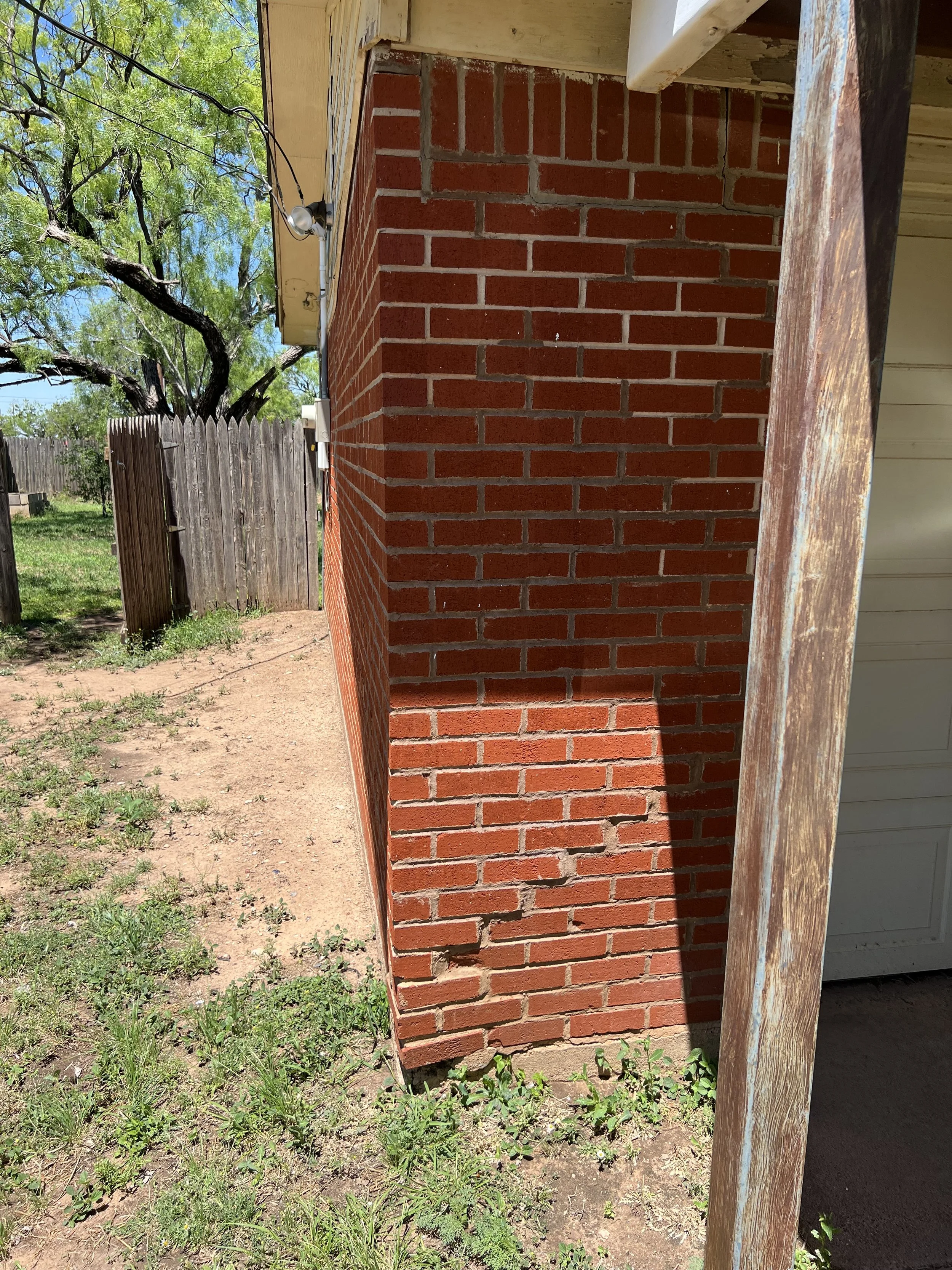 Image showing a brick wall on the outside of a house, with a wooden fence and a tree in the background. The house has a beige overhang, and part of an open garage door is visible on the right.