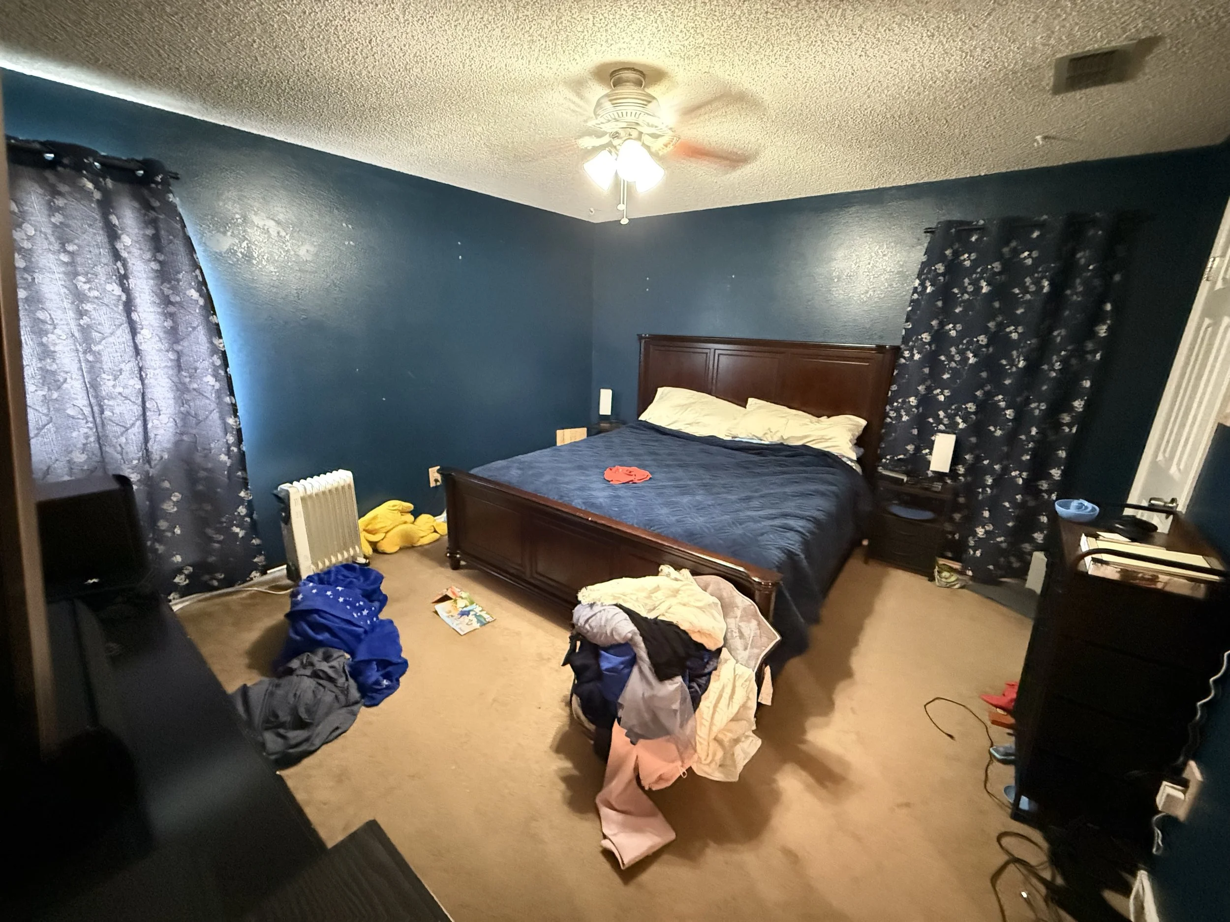 A cluttered bedroom with blue walls, a wooden bed with dark bedding, and two windows with floral curtains. The room has clothes and miscellaneous items piled on the floor and furniture.