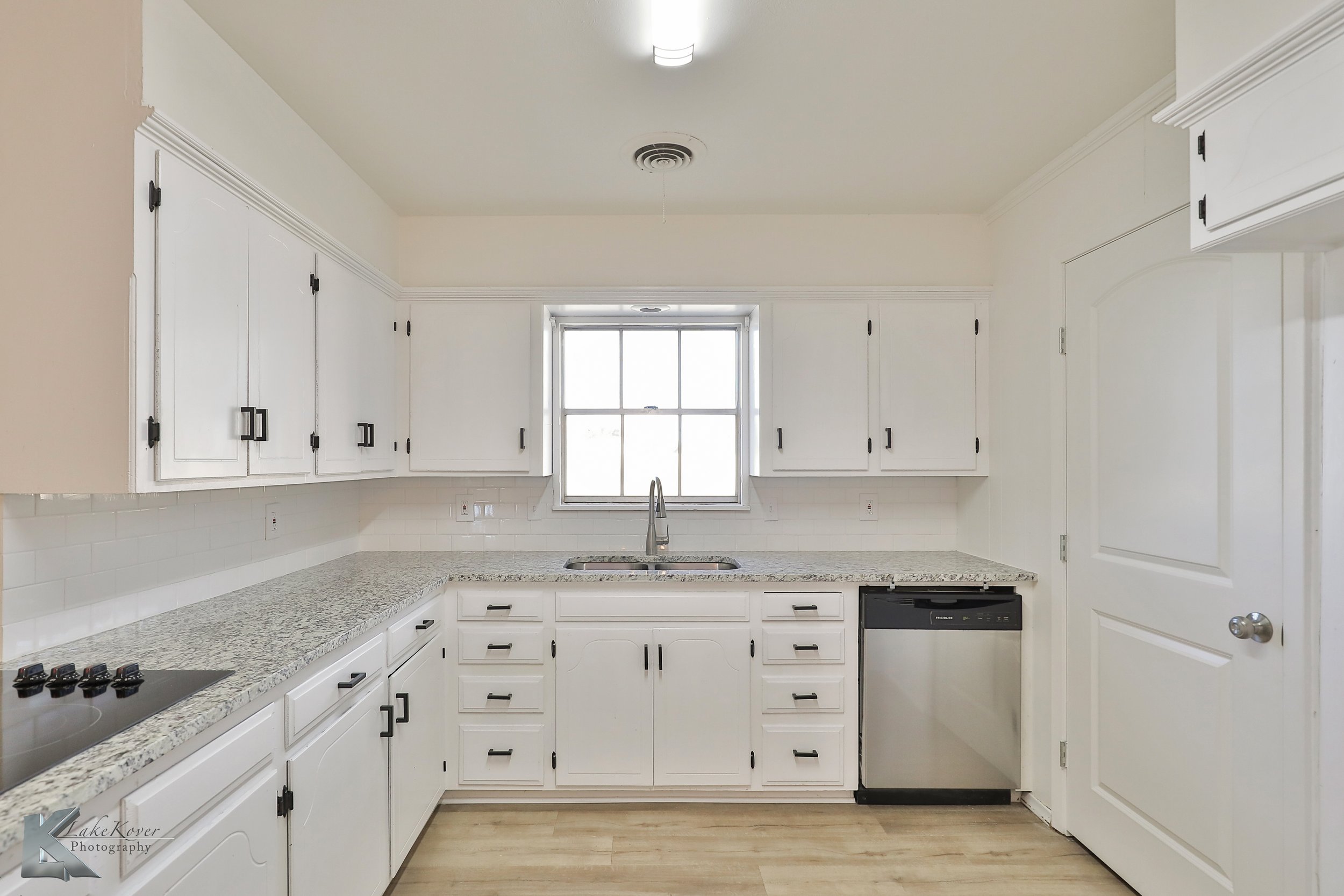 White kitchen with granite countertops, upper and lower cabinets, a window above the sink, and a dishwasher. The walls are painted white, and the flooring is light-colored wood.