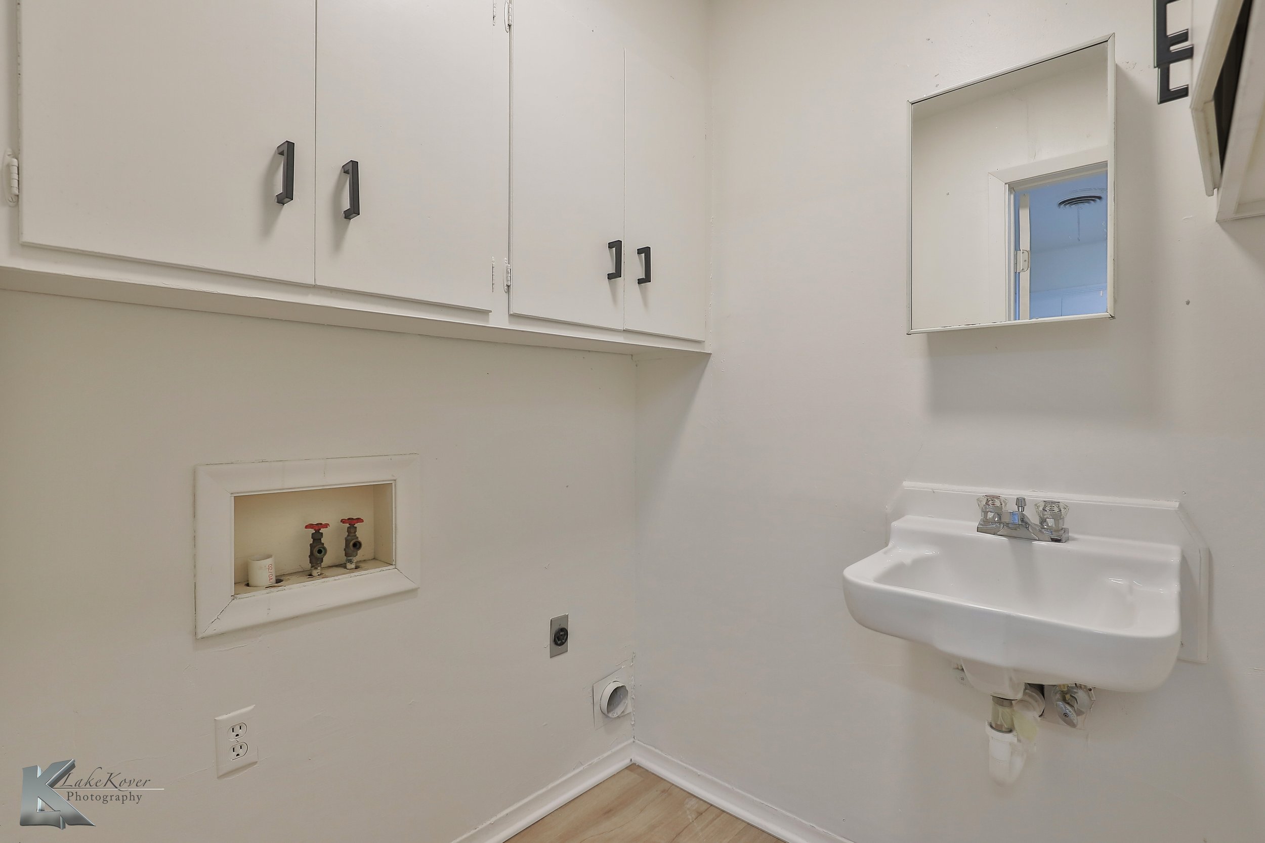 Empty laundry room with white cabinets, small sink, mirror, and exposed plumbing for washer and dryer.