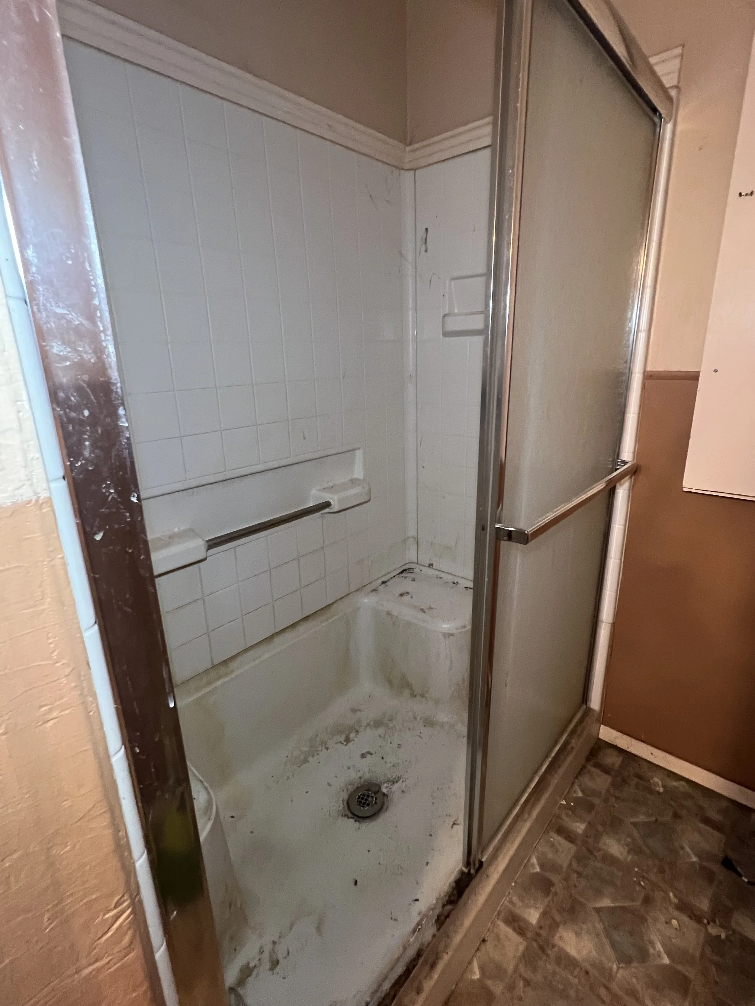 Old, dirty shower with a sliding door, white tiled walls, a grab bar, and stained floor.