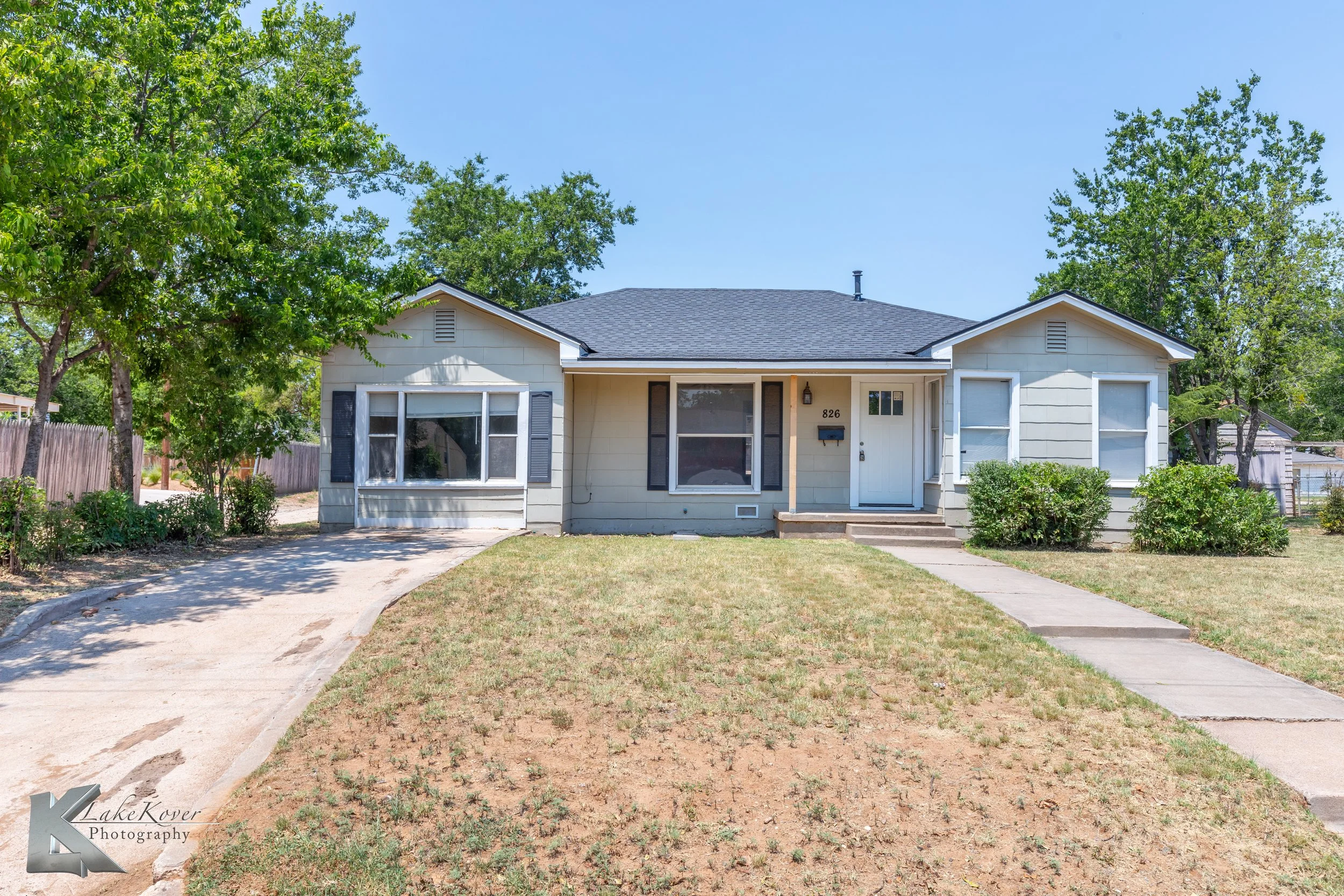 Front view of a single-story house with a paved pathway and lawn, two small bushes on each side of the front stairs, large trees in the yard, and a blue sky.