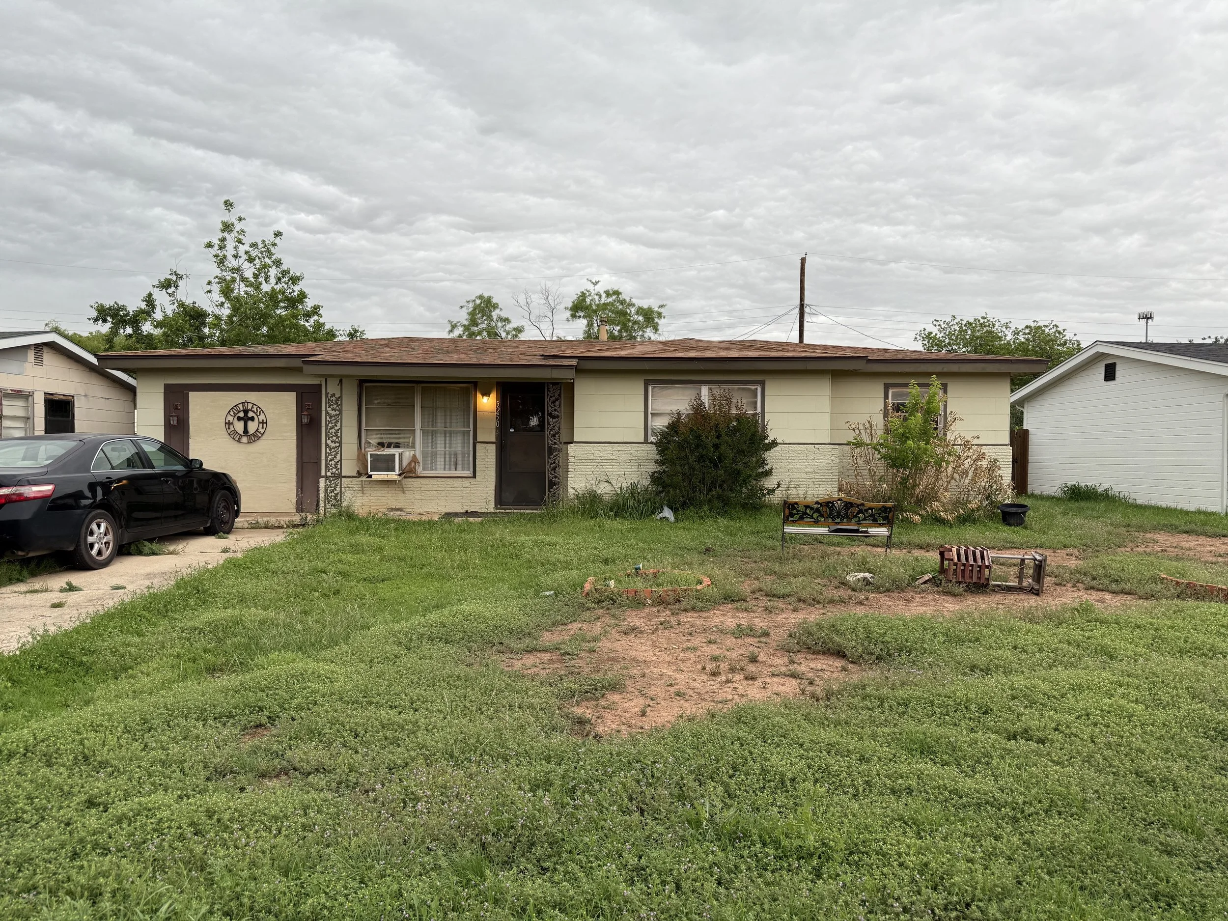 A single-story house with beige and green exterior, a small front yard with patchy lawn, a black metal bench, scattered garden decor, a black car parked in the driveway, and a cloudy sky overhead.