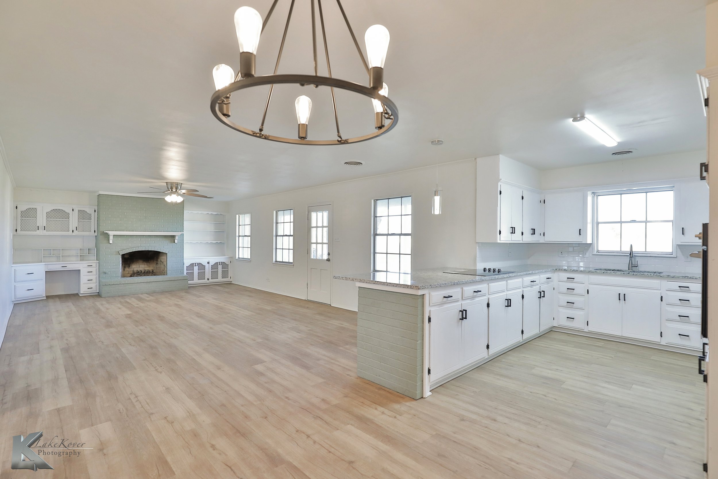 Open concept living room and kitchen with white cabinets, light wood flooring, a fireplace, multiple windows, a chandelier, and a ceiling fan.