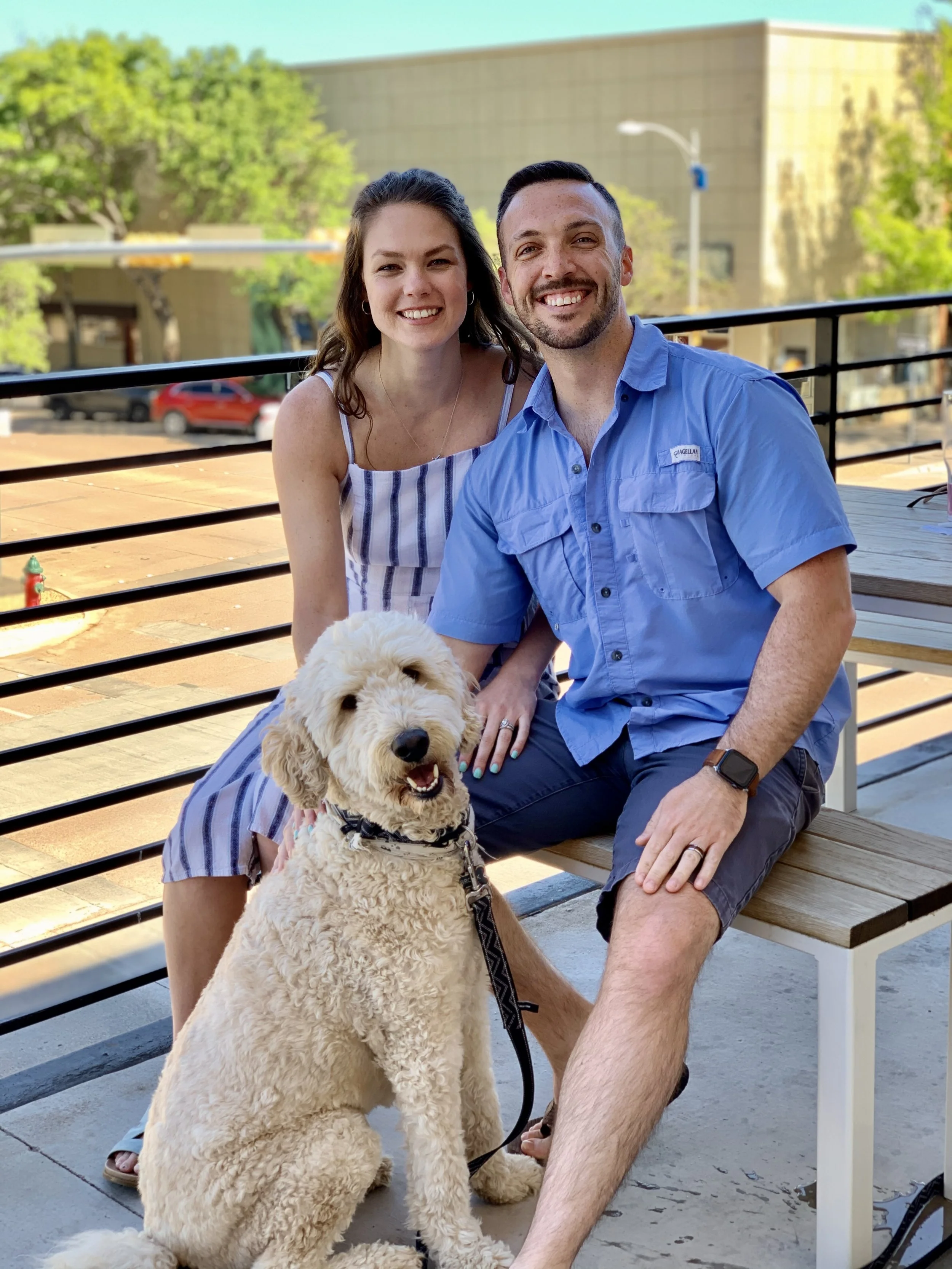 A smiling couple, a woman with long brown hair wearing a striped dress and a man with short dark hair in a blue shirt, sitting outdoors on a wooden bench with their light-colored, curly-haired dog, on a patio with a metal railing, trees, a building, and a red car in the background.