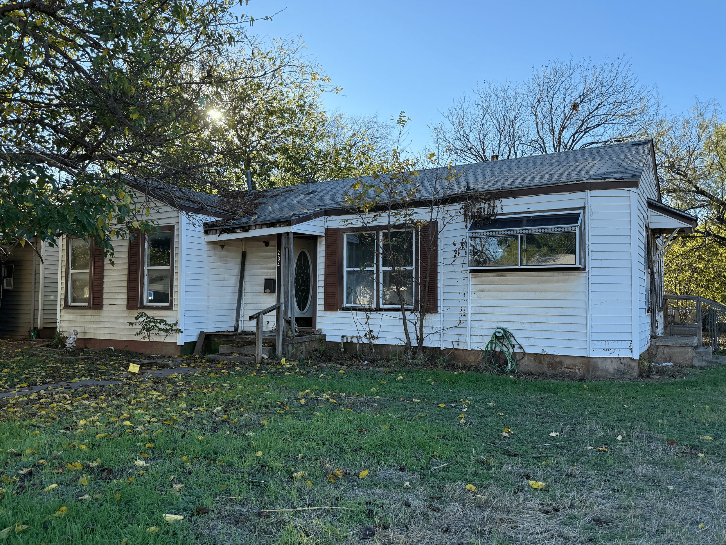 Front view of a small, older white house with brown shutters, a front porch with steps, and a sloped roof, surrounded by trees, some with bare branches, and a lawn with fallen leaves.