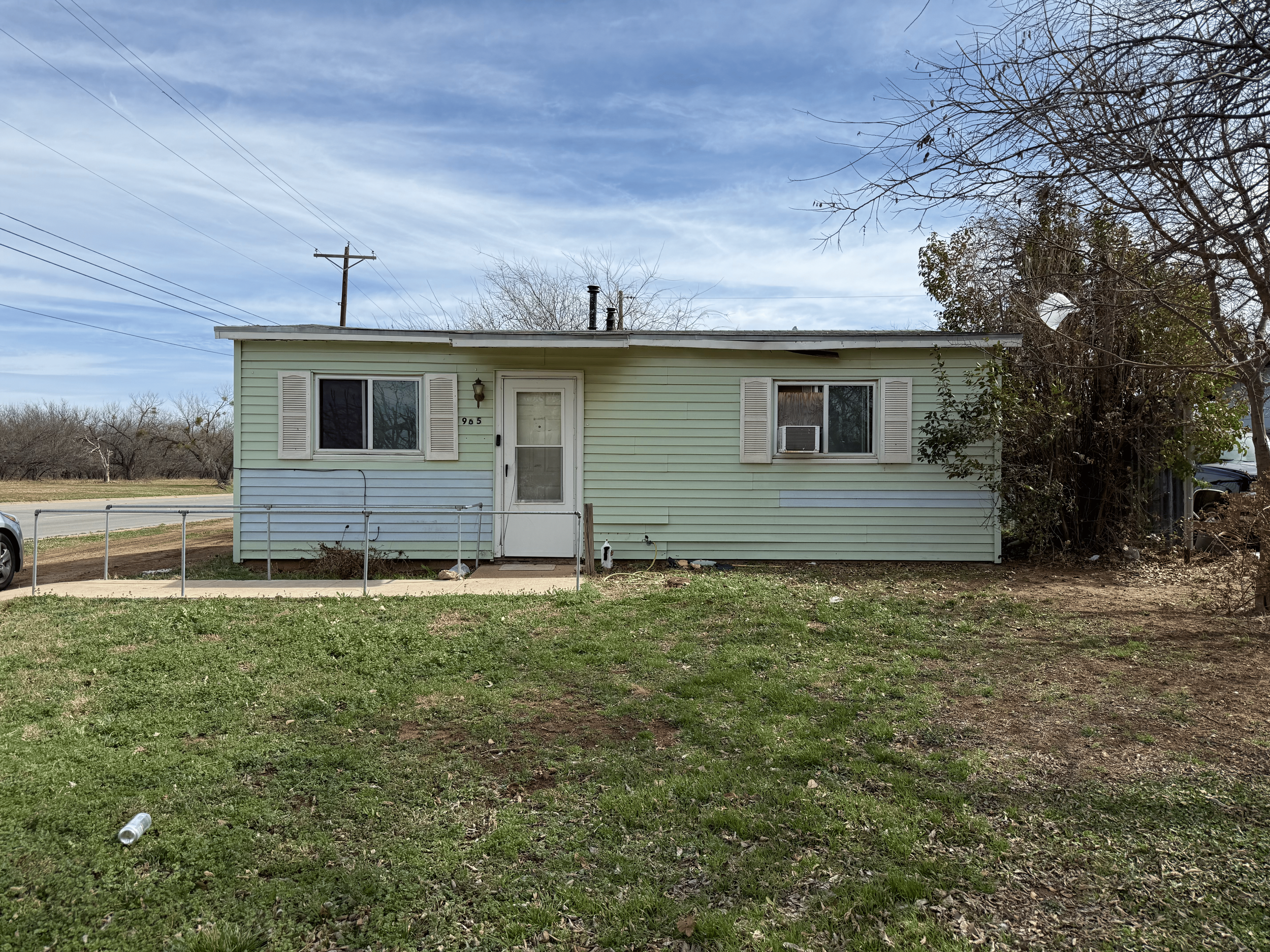 A small, light green single-story house with white shutters and a small front porch, located on a grassy yard with a sidewalk, surrounded by bare trees, under a cloudy blue sky.