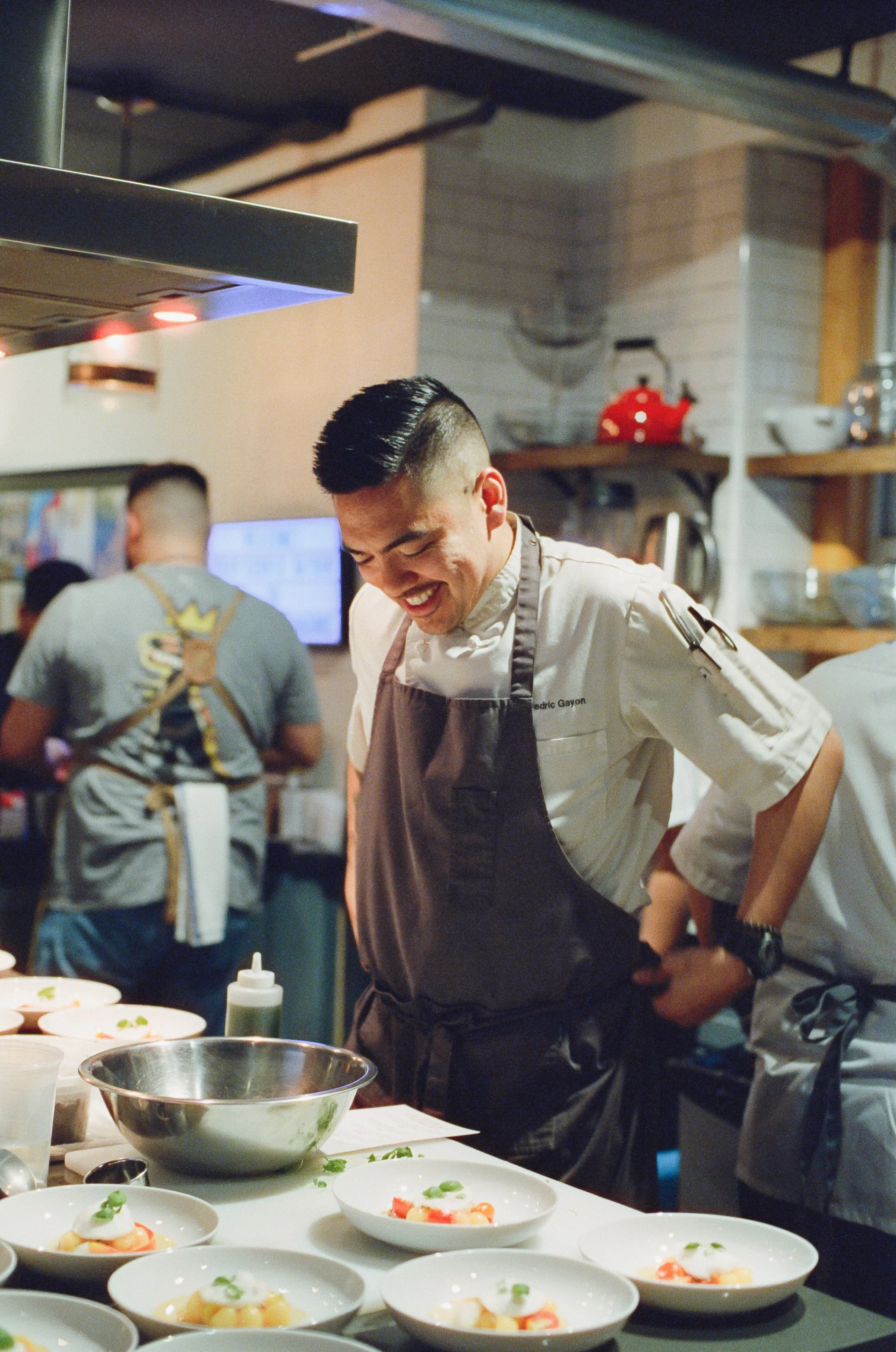 A smiling chef in a white uniform and gray apron preparing dishes in a busy kitchen with plated food on the counter. Proud of his work just like people are proud of their work.
