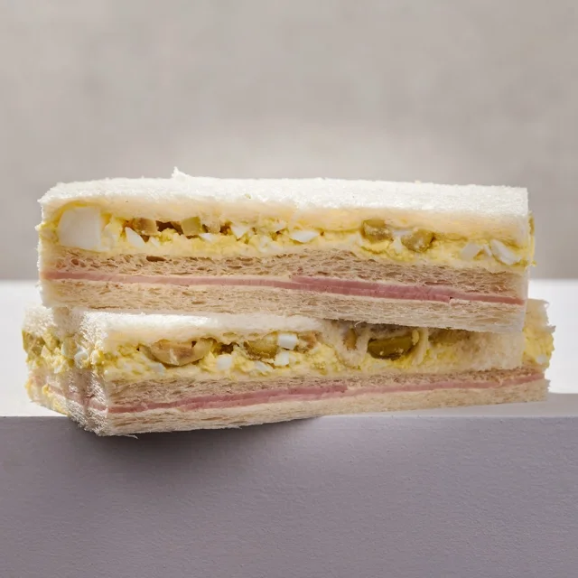 Cross-section of a layered sandwich with white bread, egg salad with chopped eggs and mayonnaise, slices of ham, and green olives.