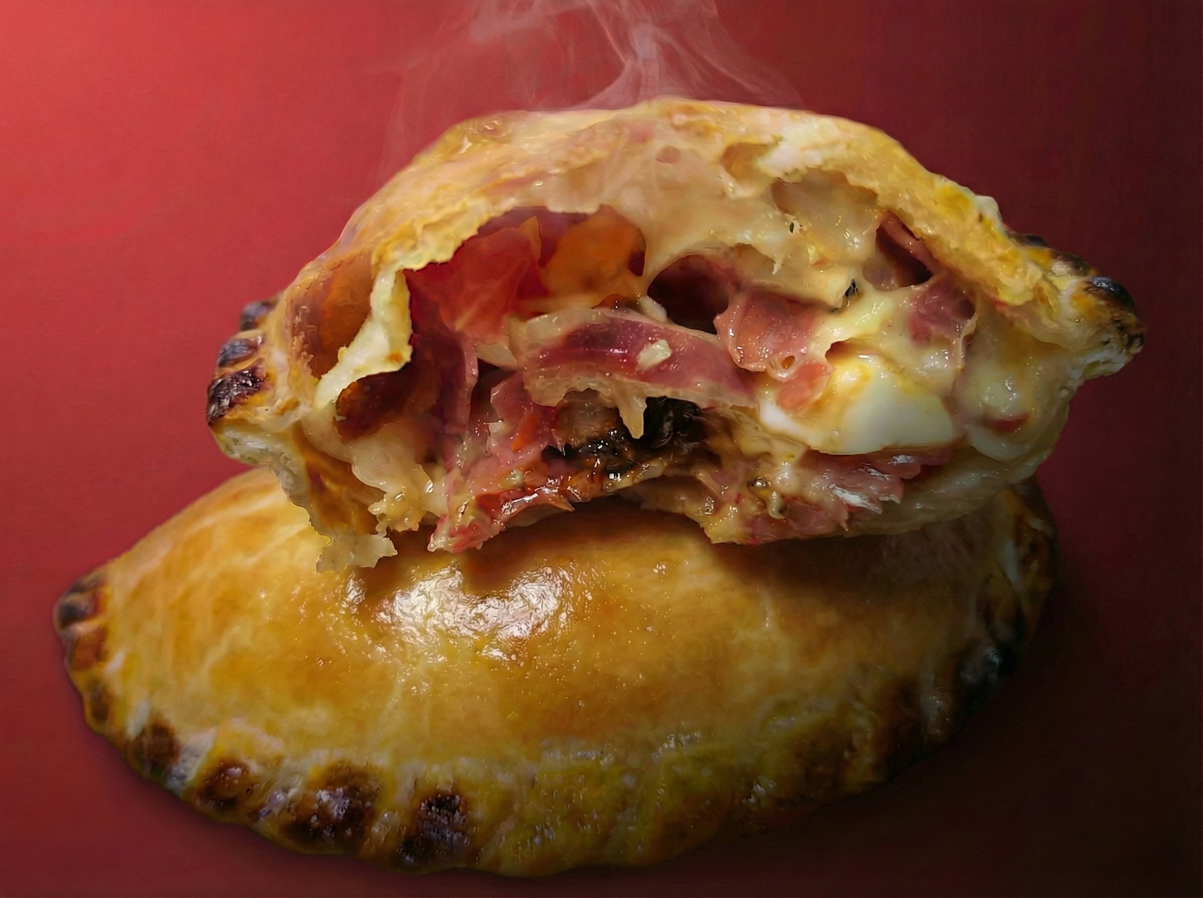 A close-up of a savory pastry with melted cheese, ham, and other ingredients, with steam rising from it, set against a red background.