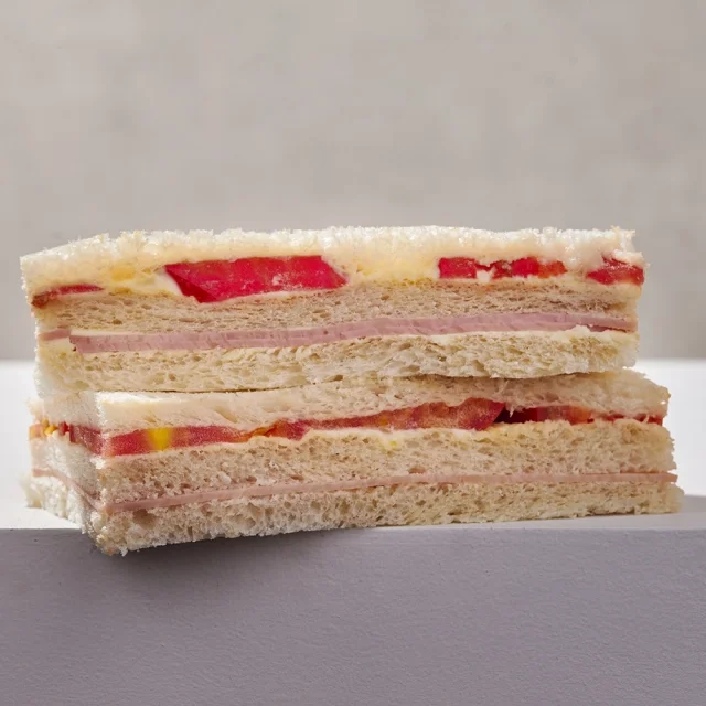 Close-up of a multi-layered sandwich with white bread, ham, cheese, tomato, and possibly mayonnaise or butter, on a white surface against a light gray background.