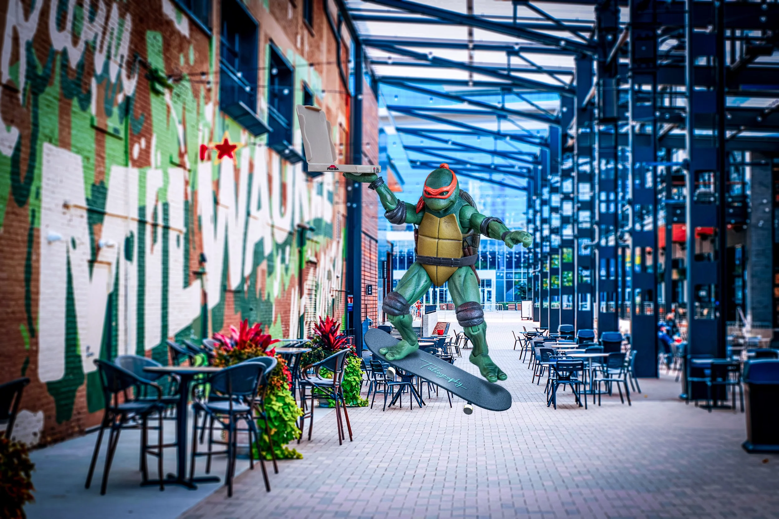 A statue of a Teenage Mutant Ninja Turtle riding a skateboard, holding a pizza box, in an outdoor seating area with tables and chairs, graffiti on a brick wall, and a modern metal structure overhead.