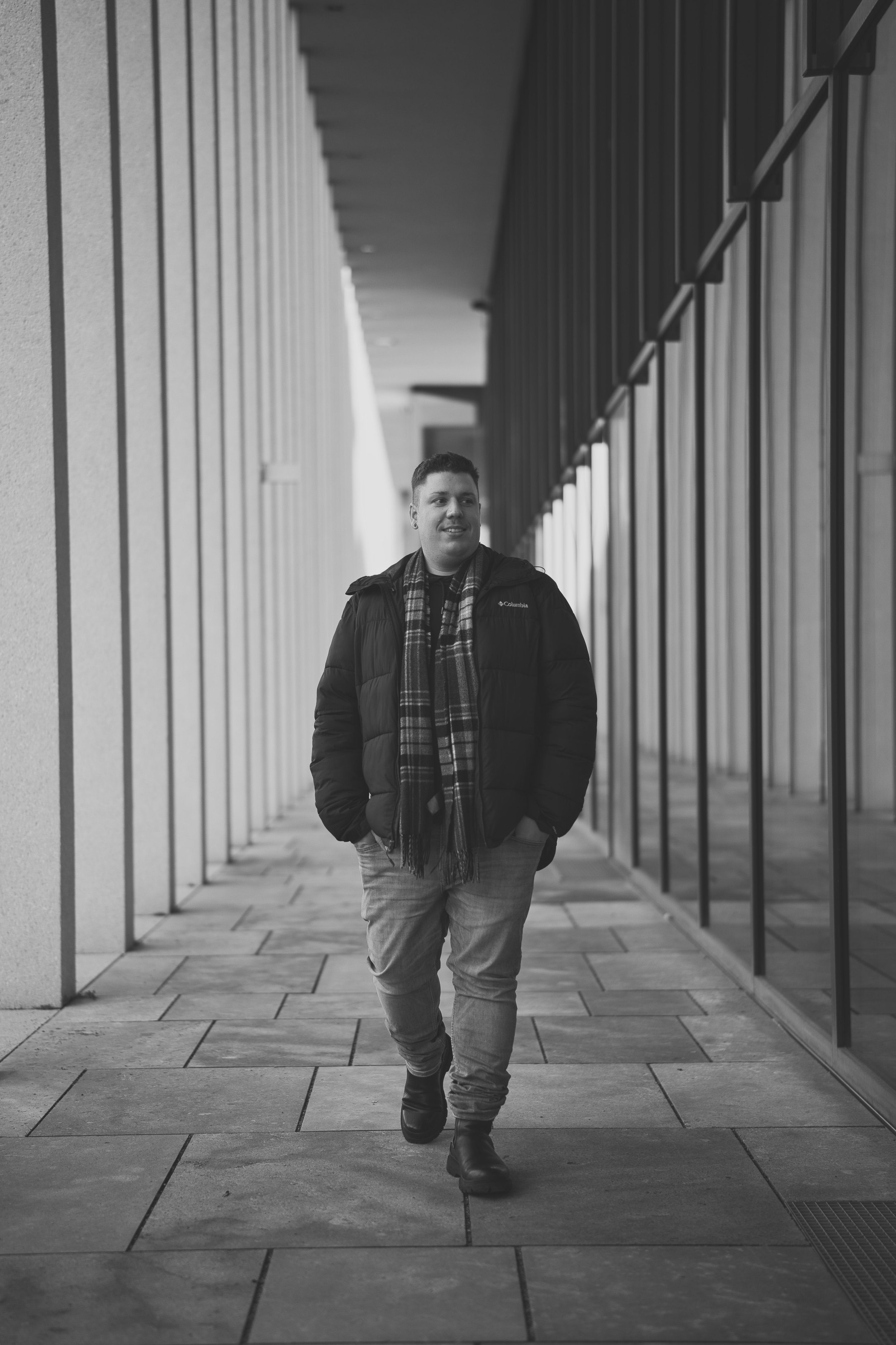A portrait of a man walking along a modern building corridor with glass windows and a concrete wall, wearing a puffy jacket, scarf, jeans, and boots.