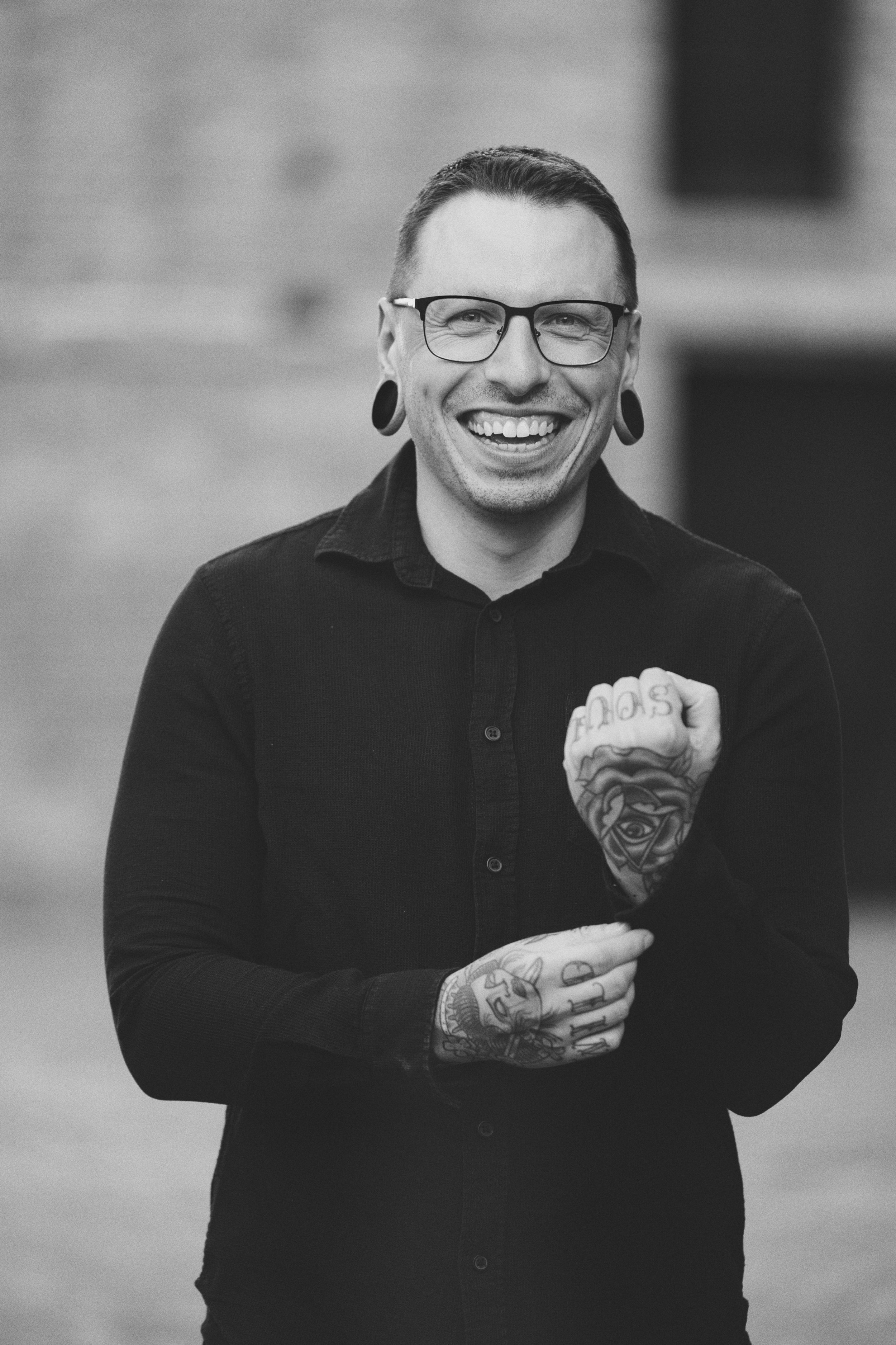 A portrait of a smiling man with glasses and tattoos on his hands, wearing a black long-sleeve shirt, standing outdoors.