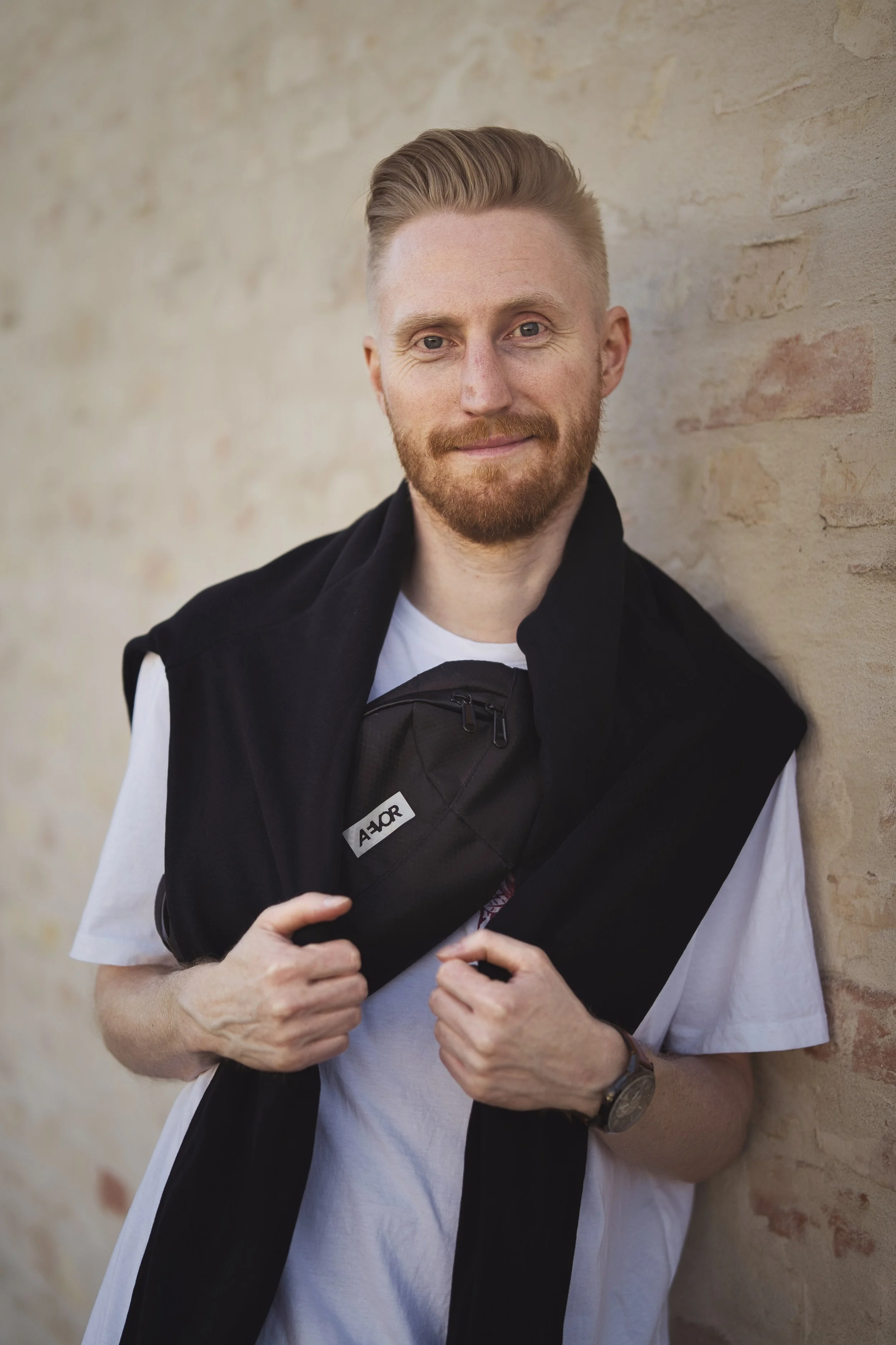 A young man with red hair and beard, wearing a white t-shirt and a black vest, standing against a brick wall. He is holding a black bag with the brand JMOR on it, near his chest, and wears a watch on his left wrist.