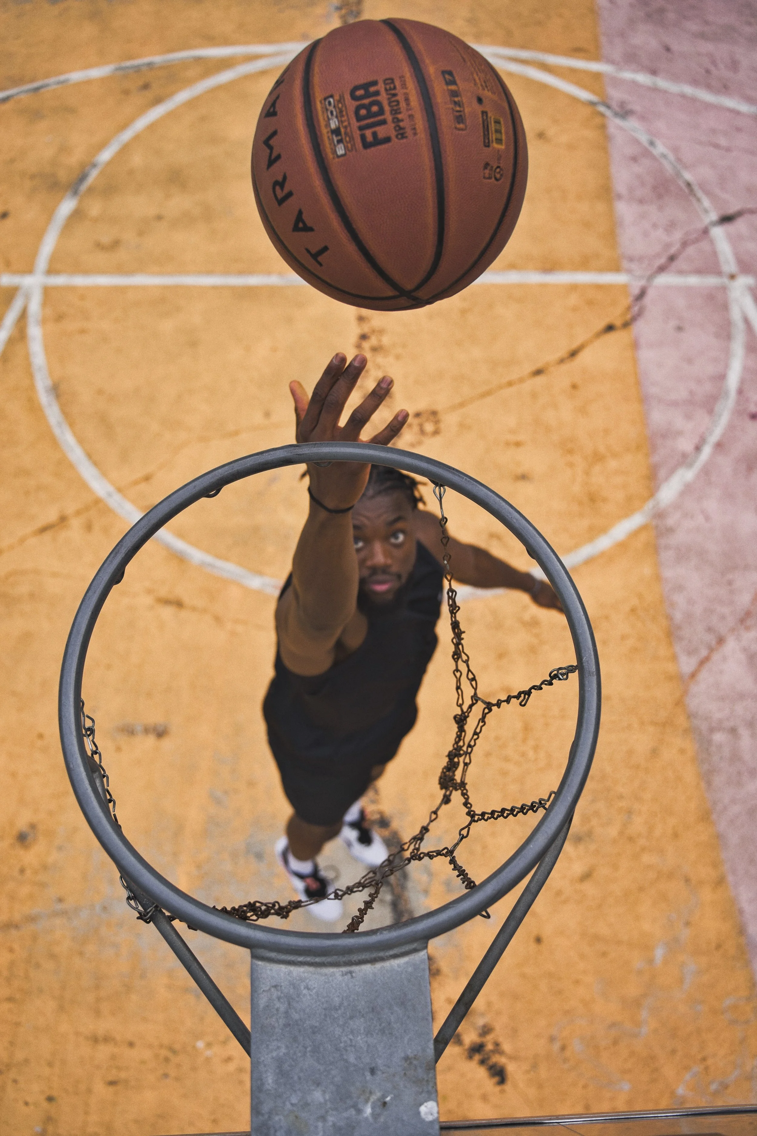 A portrait of a person reaching up towards a basketball hoop, viewed from above. The basketball is falling towards the hoop, and the person is about to catch or deflect it. The court surface is orange with white lines.