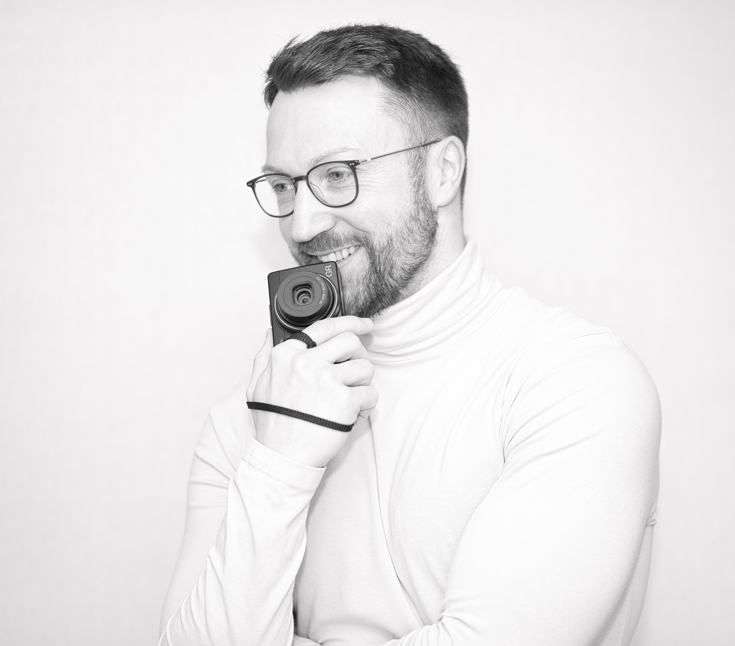 Black-and-white photo of a man with glasses, smiling, holding a camera close to his face, wearing a long sleeve shirt, against a plain light background.