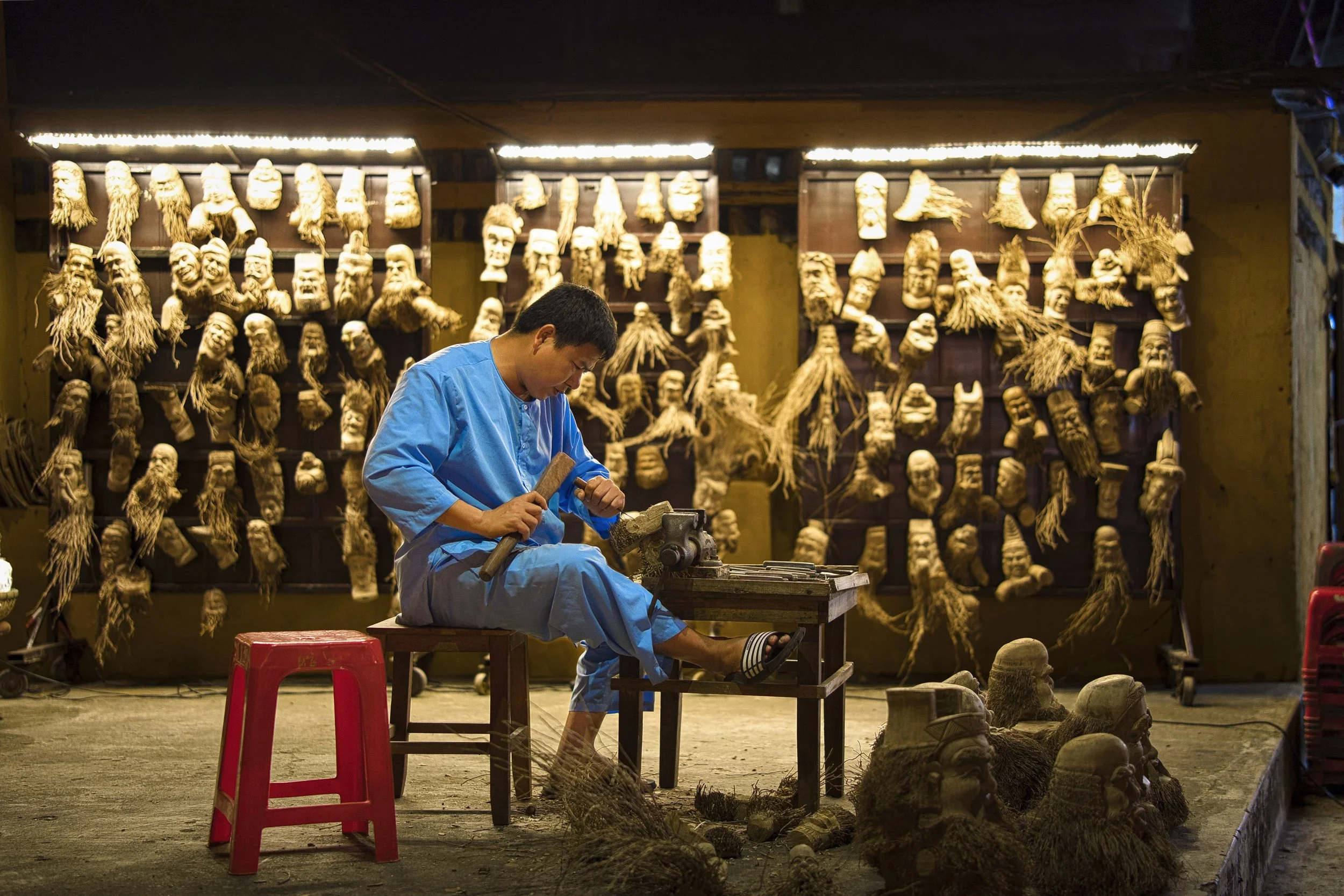 A portrait of a man working on wooden carving in front of wall decorated with various carved faces and figures in Vietnam, Hoy An.
