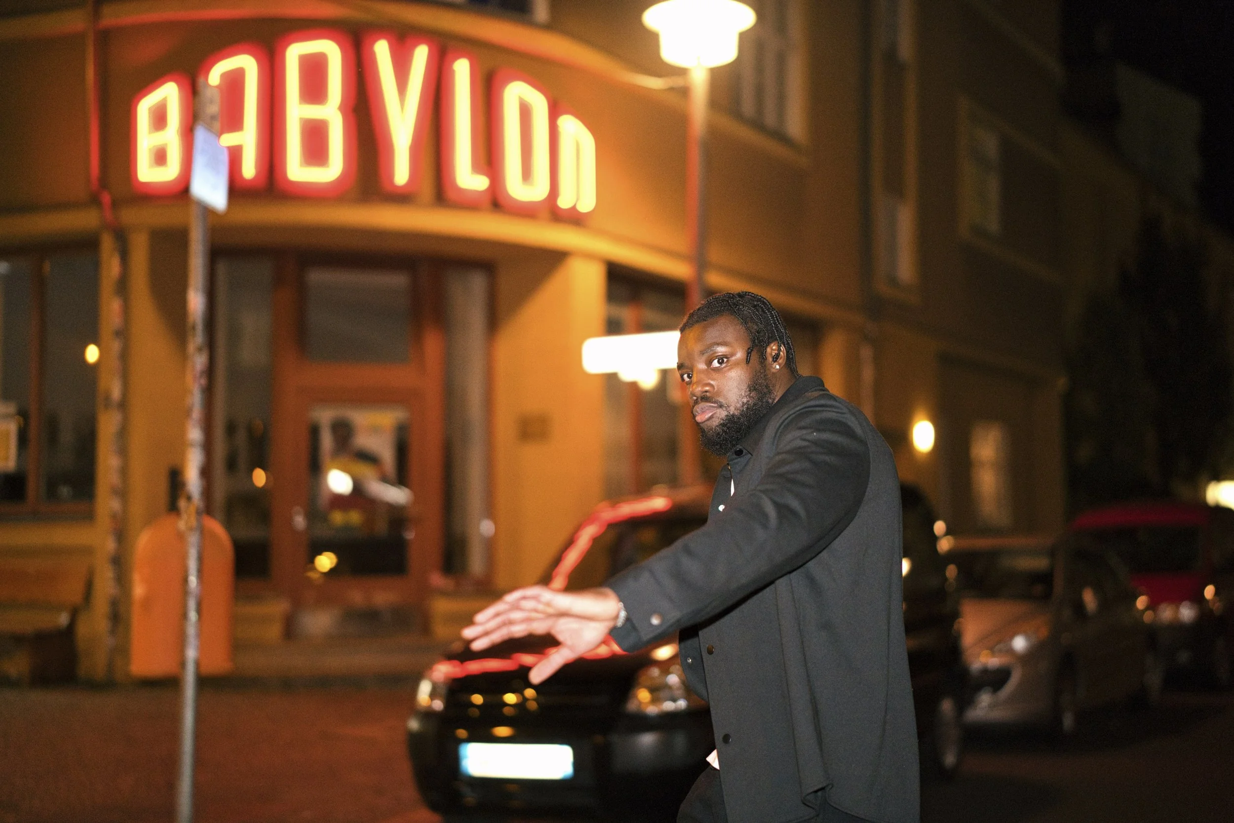A portrait of a man in Berlin with dreadlocks and a beard, wearing a black jacket, playing a keyboard on a city street at night. A neon sign reading 'Babylon' is illuminated in the background.