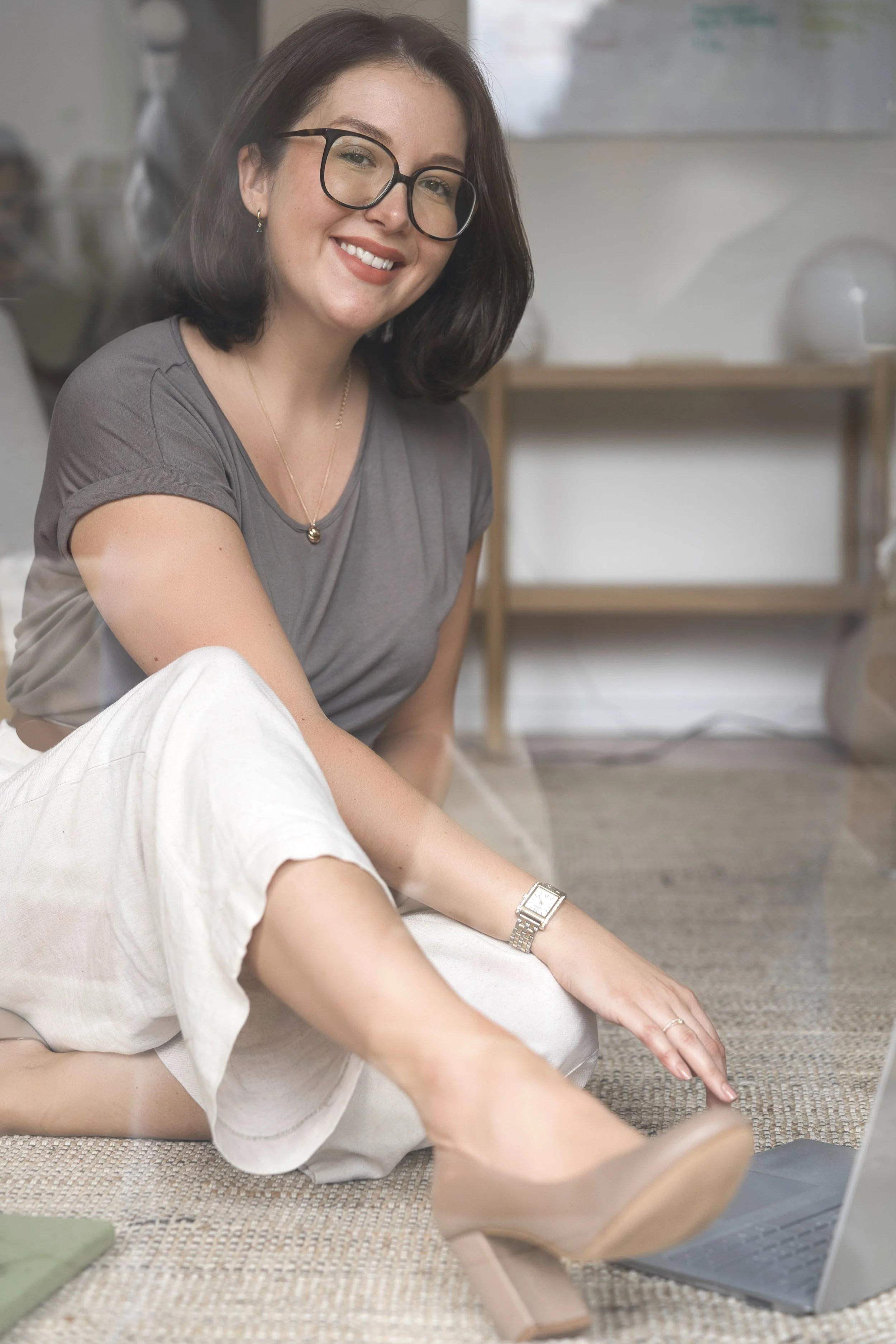 A woman wearing glasses and a gray t-shirt is sitting on the floor, smiling at the camera. She has shoulder-length dark hair, earrings, a necklace, a watch, and rings. A person in white pants is sitting beside her, with their arm pointing toward a laptop on the floor. In the background, there is a small wooden table, a white lamp, and a gray wall with a whiteboard.